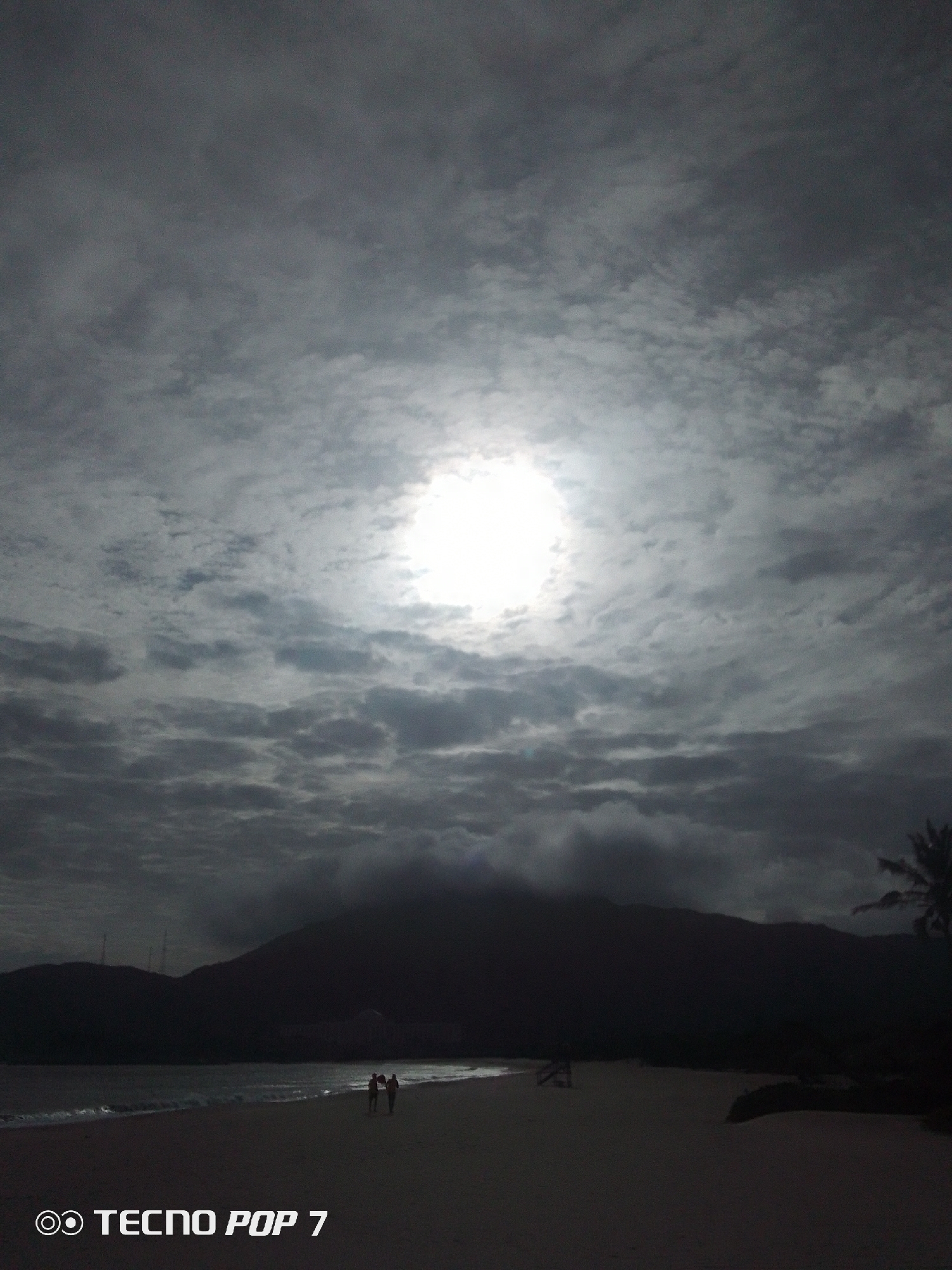 The image captures a dark beach scene in Pedro González, Venezuela. The sky is filled with dense, gray clouds, and the sun is visible through a break in the clouds. In the distance, a dark, mountainous landmass rises from the horizon. The beach itself appears wide and sandy. Two people can be seen walking along the shoreline. A palm tree is present in the foreground on the right side of the image. The image is underexposed, giving it a somber, almost monochromatic quality. "Tecno Pop 7" is printed on the lower left-hand corner of the image.