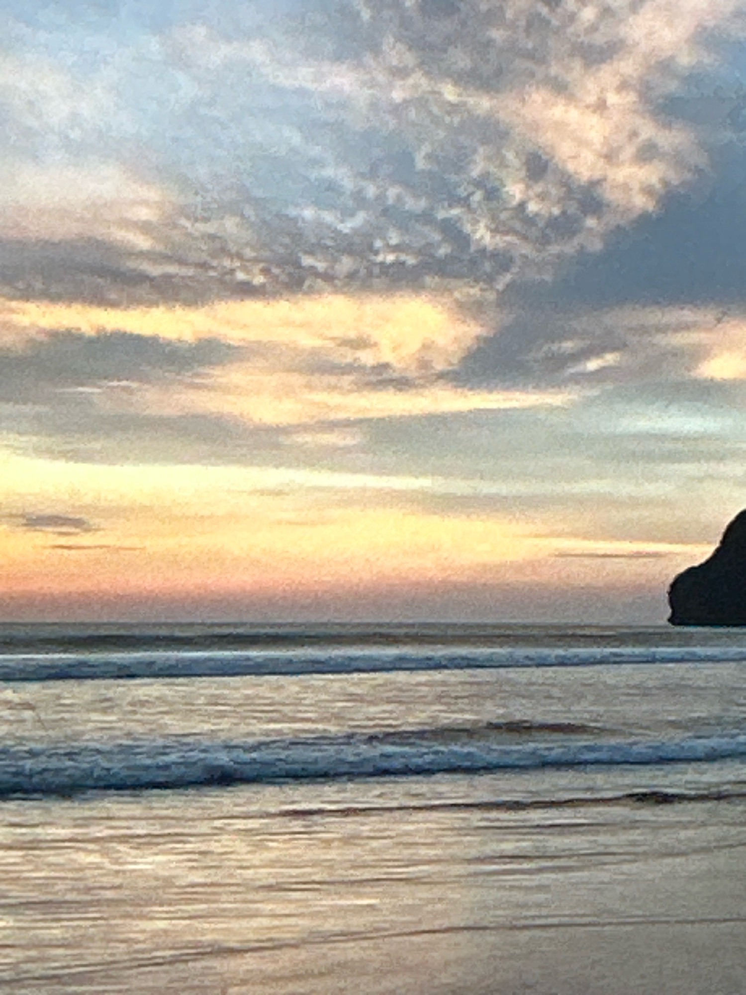 The image captures a serene beach scene at dusk. The sky is a canvas of soft, diffused colors, with streaks of pale yellow and pink blending into a muted blue. Wispy clouds are scattered across the sky, catching the last rays of sunlight. The ocean is calm, with gentle waves rolling towards the shore. Three distinct lines of waves are visible, each with a subtle white crest. The wet sand in the foreground reflects the warm tones of the sky. On the right side of the horizon, a dark, silhouetted rock formation juts out from the water. The overall impression is one of tranquility and natural beauty.