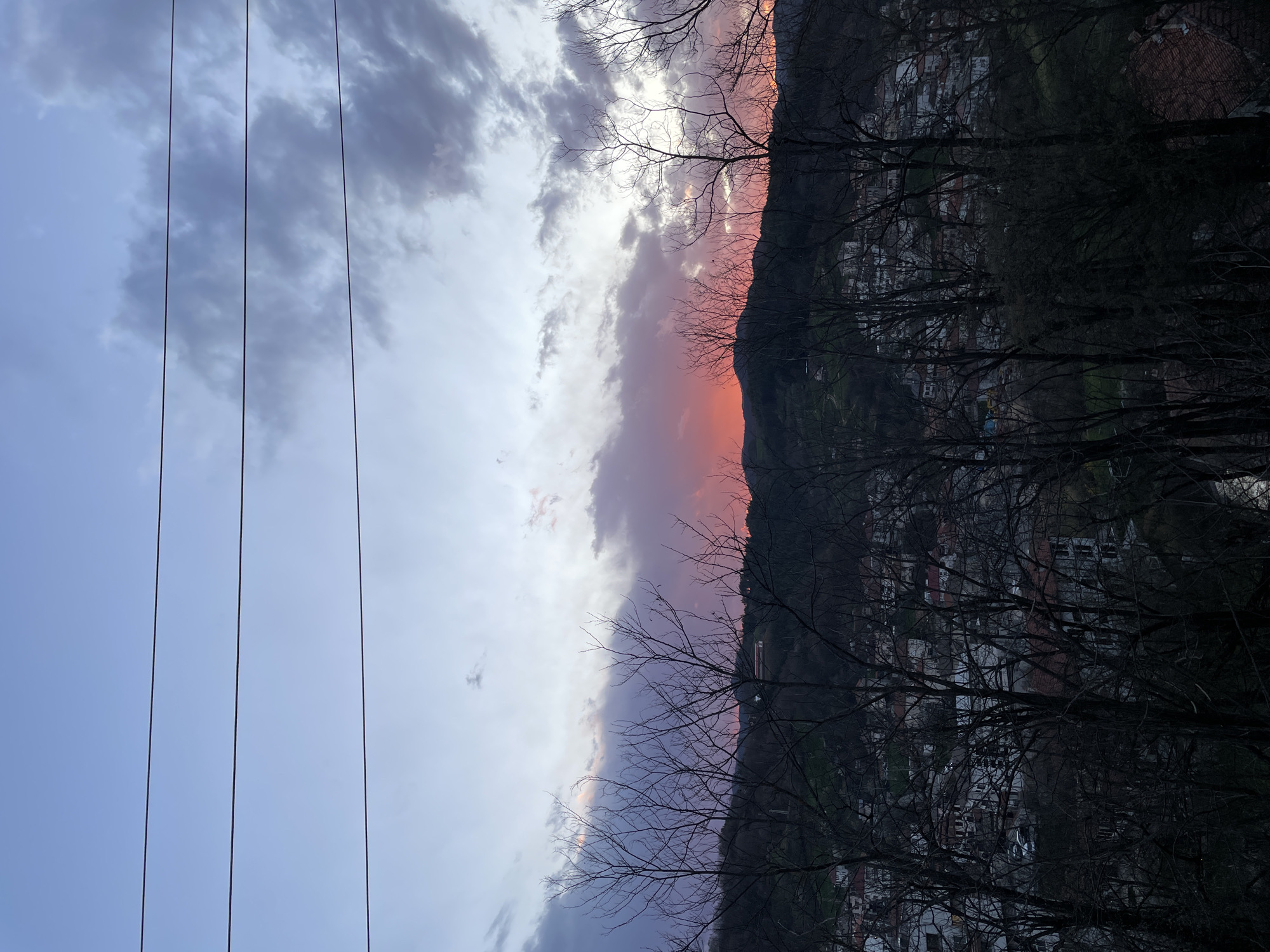 The image depicts a twilight scene. The sky transitions from a pale blue on the left to clouds with orange and pink hues towards the center, likely reflecting the setting sun. Power lines are visible against the pale blue sky.
On the right side of the image is a dark mountain range with buildings clustered on its lower slopes. Bare tree branches are silhouetted against the sky and mountains. Given the context, the location is likely in or around Zlatograd, Bulgaria, a town situated in a mountainous region.