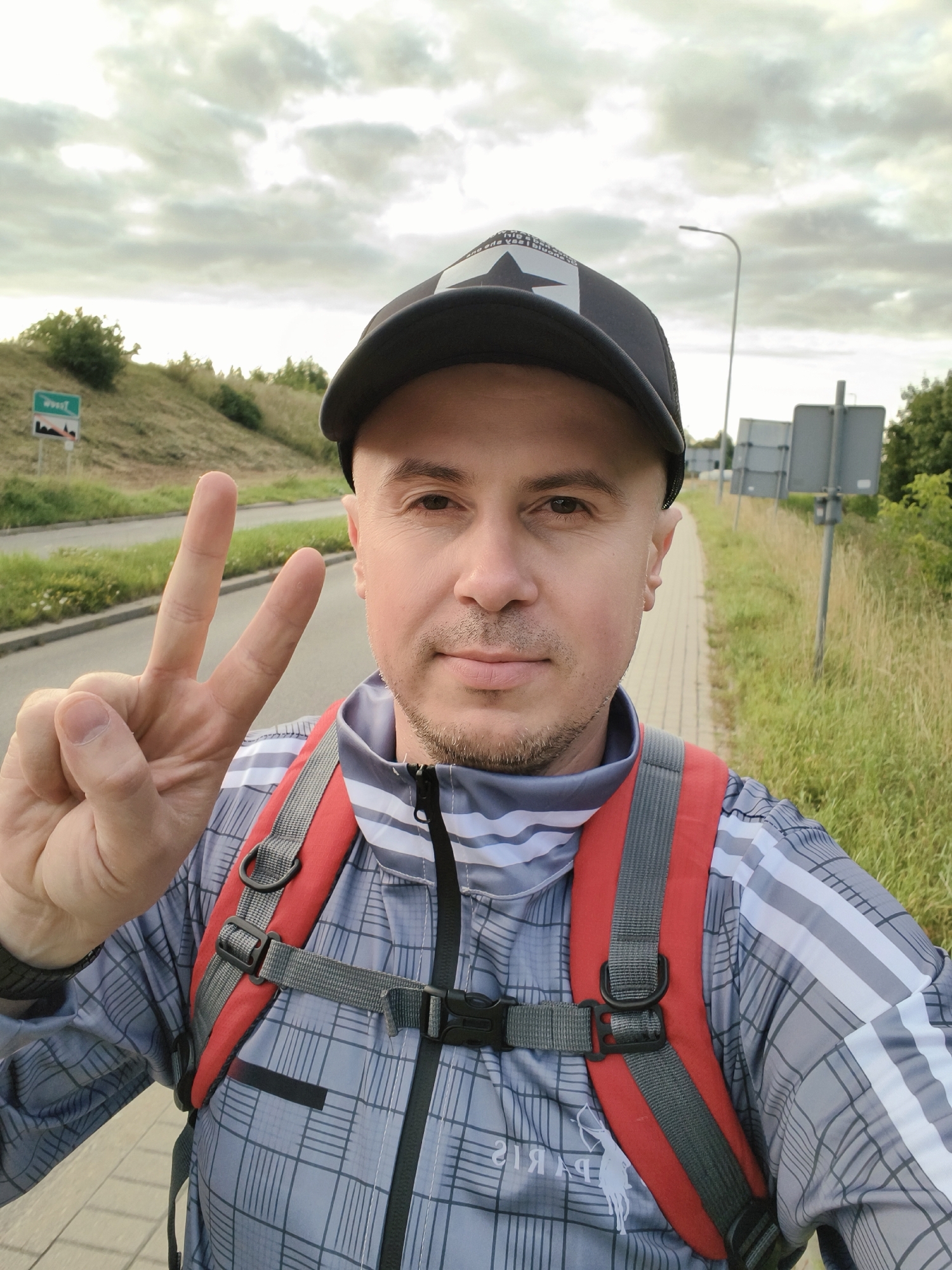 A man in a grey checkered jacket and a black baseball cap is posing for a selfie on a paved sidewalk next to a road. He is holding up his left hand with his index and middle fingers extended in a peace sign. He is wearing a red and grey backpack. The background shows a grassy embankment with a road, a street light, and some trees. There are also traffic signs visible in the distance. The sky is cloudy. The location appears to be on the outskirts of Tczew, Poland, given the presence of a sign with Polish text and the overall road and landscaping characteristics common in the region.