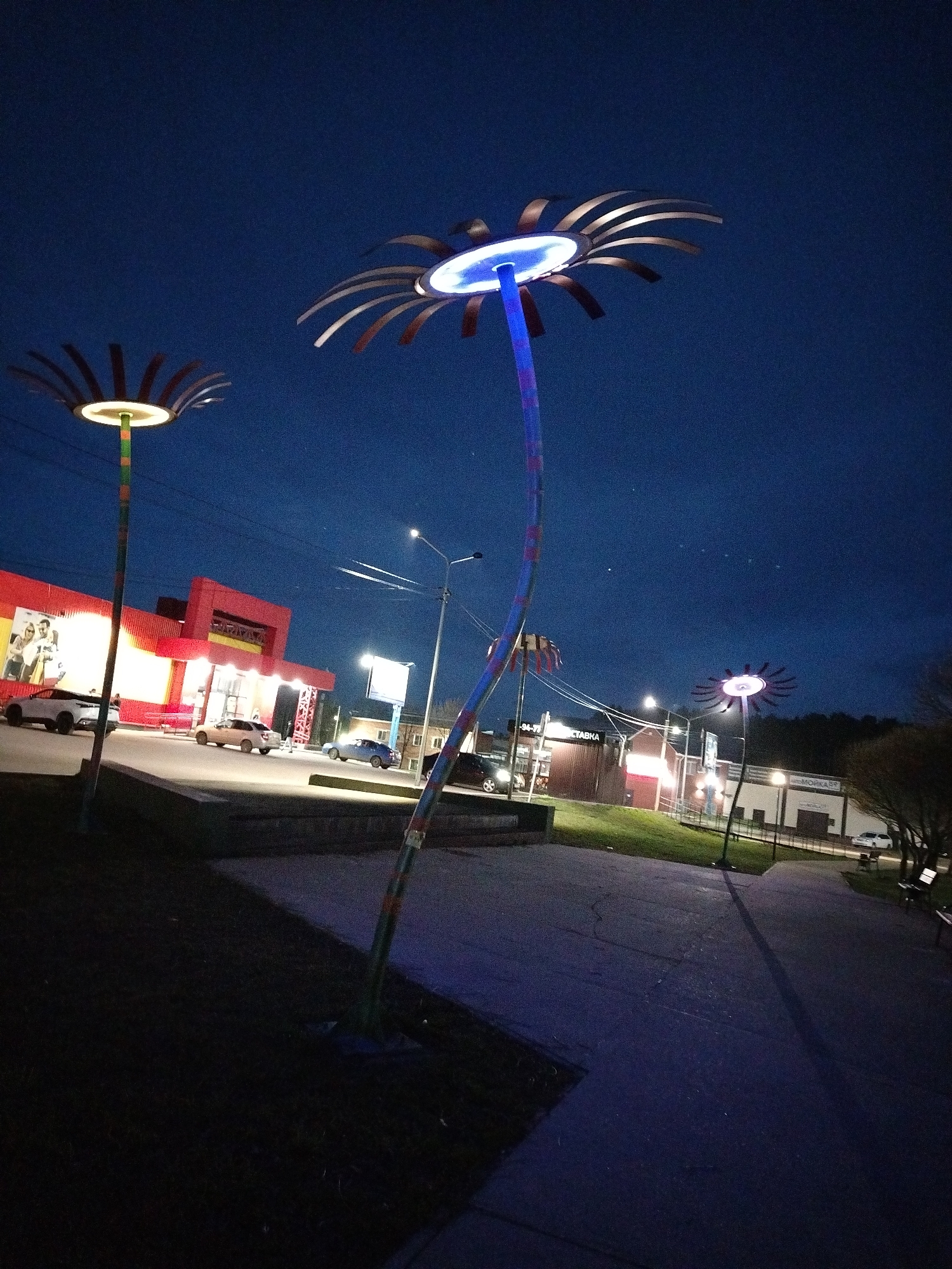 The image captures an evening scene in Chaikovsky, Russia, featuring a public area illuminated by unique, flower-shaped streetlights. The time is dusk or night, indicated by the dark blue sky and the bright artificial lights.
In the foreground, a paved walkway or path stretches into the distance, with patches of dark grass on either side. Three prominent streetlights are visible, designed to resemble flowers with radiating petals. The closest and tallest streetlight, centrally located, has a tall, curved stem and emits a bright blue light from its center. To its left, another similar streetlight has a straighter stem and glows with a warm yellow-white light. A third, smaller flower-shaped light can be seen further down the path on the right, emitting a reddish-purple light.
Behind the foreground elements, a street with several cars is visible. A large, brightly lit red building with yellow accents dominates the left side of the midground, appearing to be a commercial establishment, possibly a supermarket. Further back on the right, other commercial buildings with various facades (brown and white) can be seen. There are utility poles with overhead wires crisscrossing the sky.
Activity appears to be minimal, characteristic of an early evening in a public space. Cars are parked or driving along the street, and a few indistinct figures are near the red building.
Notable details include:
* **Time of day:** Evening or night, with a clear, deep blue sky showing a few faint stars.
* **Location cues:** The architecture and visible text suggest a Russian urban setting.
* **Visible text:** On the red building, a large advertisement poster features people, and above its entrance, a sign likely reads "ДИКСИ" (Dixy), a Russian supermarket chain. Further right, a sign on a brown building seems to say "СТАВКА" (STAVKA, meaning "bet" or "rate"), and another on a white building spells out "МОЙКА" (MOYKA, meaning "car wash"). Another sign partially visible includes "-94-77".