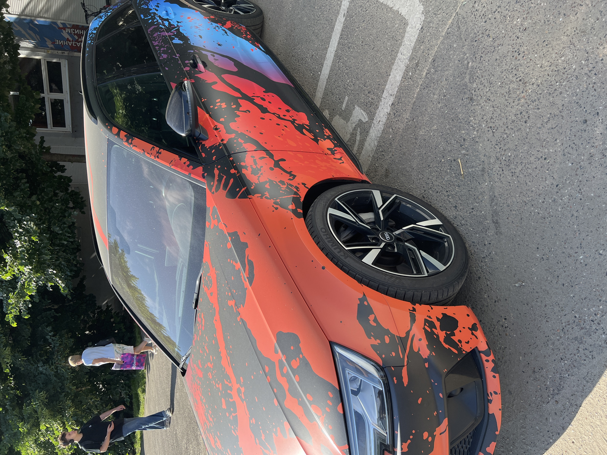 A black Audi A4 Avant station wagon with a bold red and black splatter graphic wrap is parked in a paved lot. The car is viewed from a low angle, emphasizing its front left quarter. The graphic extends from the hood, across the fenders, and onto the side of the vehicle, transitioning into blue and pink hues towards the rear. Two individuals are visible in the background on the left. One, a young man wearing a black t-shirt and blue jeans, walks away from the camera. The other, an older woman in a light blue t-shirt and patterned shorts, walks towards the camera carrying a purple shopping bag. In the far background, a building with large windows and a tree with green foliage are visible. The scene appears to be an outdoor environment, possibly a parking lot in a city. The car's license plate is not visible. The pavement has visible white parking lines. The image is taken during daylight with sunlight casting shadows.