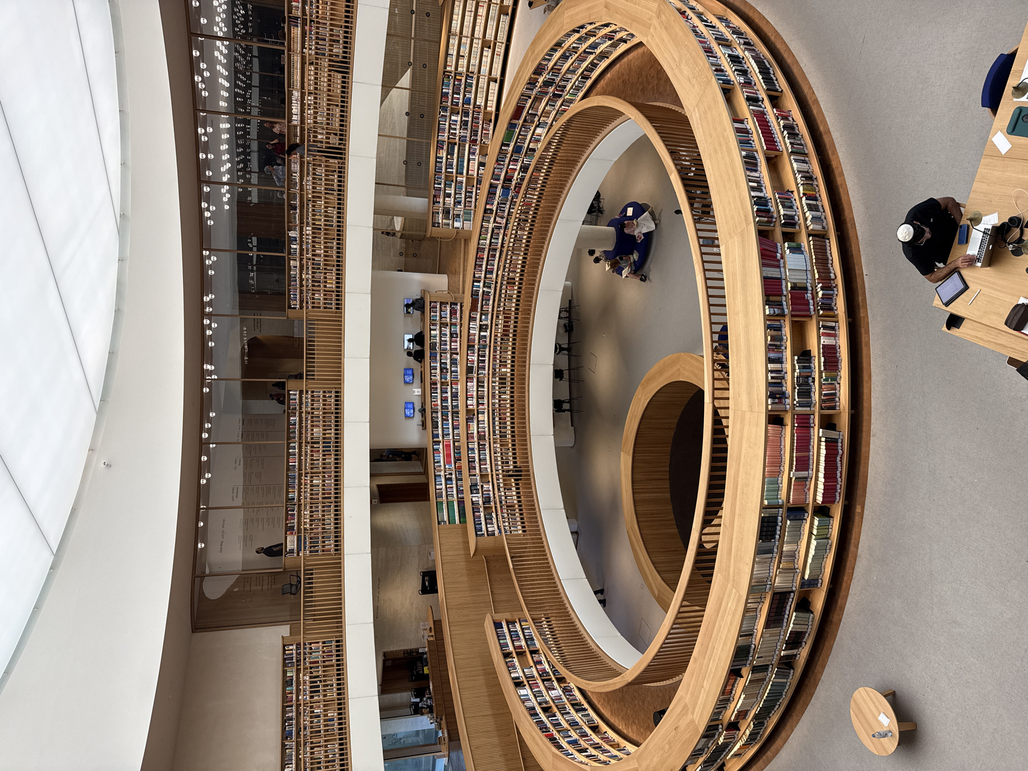 This image depicts the interior of a modern, multi-level library with a distinctive spiral design, located in Jerusalem, Israel. The library features curved wooden bookshelves that ascend in a helical fashion around a central open space. The shelves are filled with numerous books. Natural light and artificial lighting, from overhead fixtures and lamps, illuminate the space.
On the upper levels, visible through glass partitions and along open balconies, are more bookshelves and people reading or browsing. Several individuals are seated at desks with laptops, and others are gathered in conversation.
In the foreground, a man wearing a kippah is seated at a wooden table, working on a laptop. A tablet, phone, and papers are on the table. In the middle ground, a group of people are seated on a blue, curved couch in the open central area, some reading. A small, round wooden table with a bottle of water and a packet of tissues sits on the carpeted floor near the curved bookshelves.
The architecture is characterized by light wood, white walls, and a bright, diffused ceiling. The overall atmosphere is one of quiet study and intellectual engagement.