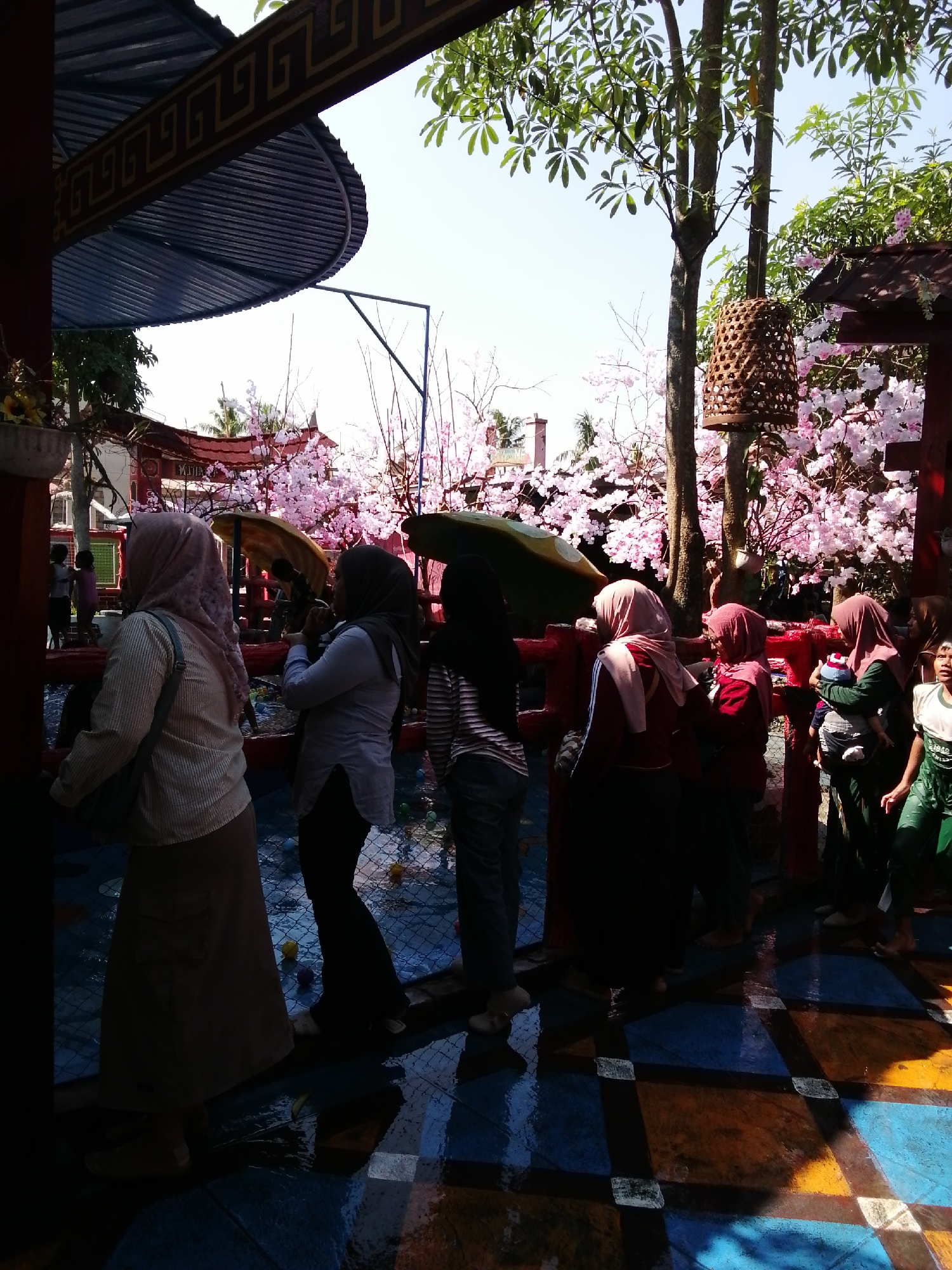 The image depicts a street scene in Tegalwangi, Indonesia, characterized by a bright, sunny sky and lush greenery. In the foreground, several individuals are observed, predominantly women wearing headscarves and modest attire. One woman in the left foreground wears a light-colored, vertically striped long-sleeved shirt and a long, light brown skirt. To her right, another woman is visible from the waist down, wearing a dark, flowing garment and dark pants. Further to the right, a woman in a light blue, long-sleeved shirt and dark pants is seen with her back to the camera.
Behind the people, a vibrant display of artificial cherry blossoms in various shades of pink adds a festive atmosphere. Red railings line the area, separating the onlookers from a colorful tiled floor, which appears wet and reflects the sunlight. Several small, round objects, possibly toys or balls, are scattered on the tiled surface.
In the background, traditional Indonesian architecture is evident, with red buildings featuring ornate details and curved roofs. A tall tree with branches laden with green leaves stands prominently, and a rustic, cylindrical woven lantern hangs from one of its branches. The overall scene suggests a public gathering or a cultural event, possibly a temple or a recreational area, given the decorative elements and the presence of children in the middle ground. The atmosphere is lively and engaging, with a blend of cultural and natural elements.