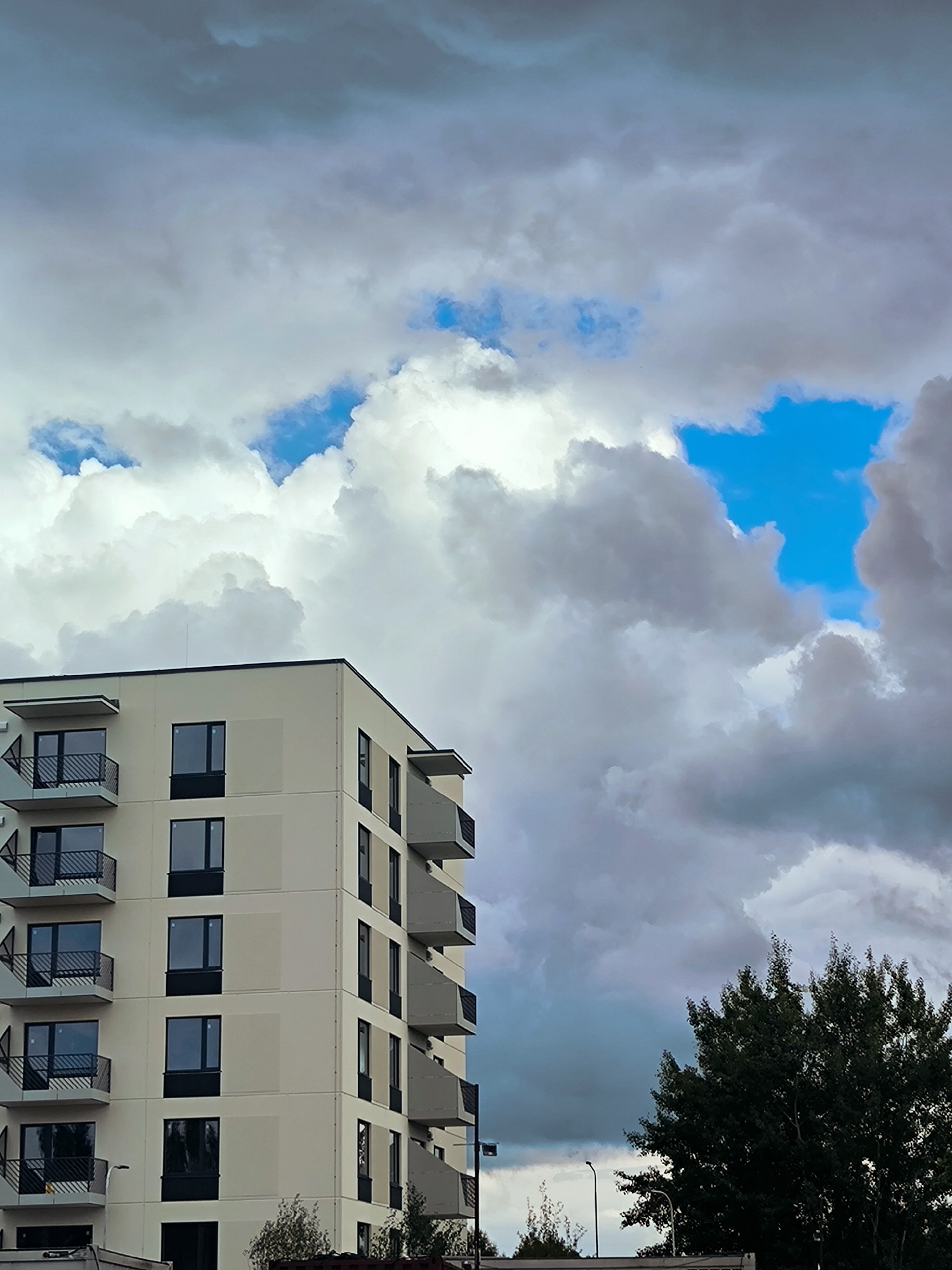 A modern, light-colored apartment building stands to the left of the frame, its balconies and dark-paned windows clearly visible. The building occupies approximately the left third of the image. To the right, a large, dark green tree fills the lower right portion of the scene. Above the building and tree, a dramatic sky is filled with a mix of heavy, dark gray clouds and bright white cumulus clouds, with patches of vivid blue sky visible through the gaps. The scene appears to be an exterior view on an overcast day, with a strong contrast between the building and the dynamic sky. The location is the city of Riga, Latvia.