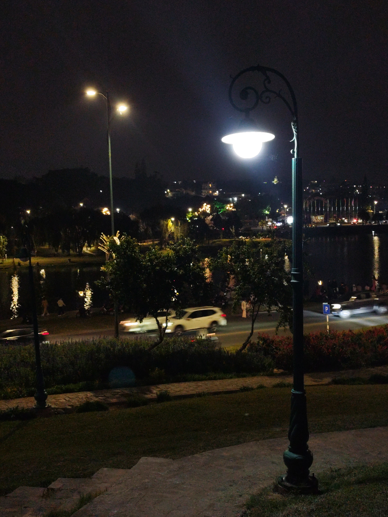 This is a nighttime scene in Da Lat, Vietnam. A decorative streetlight stands prominently in the foreground. Further back, a body of water reflects lights from the city. Cars are traveling along a road that runs parallel to the water. People are visible walking along the water's edge. Other lights illuminate the landscape.