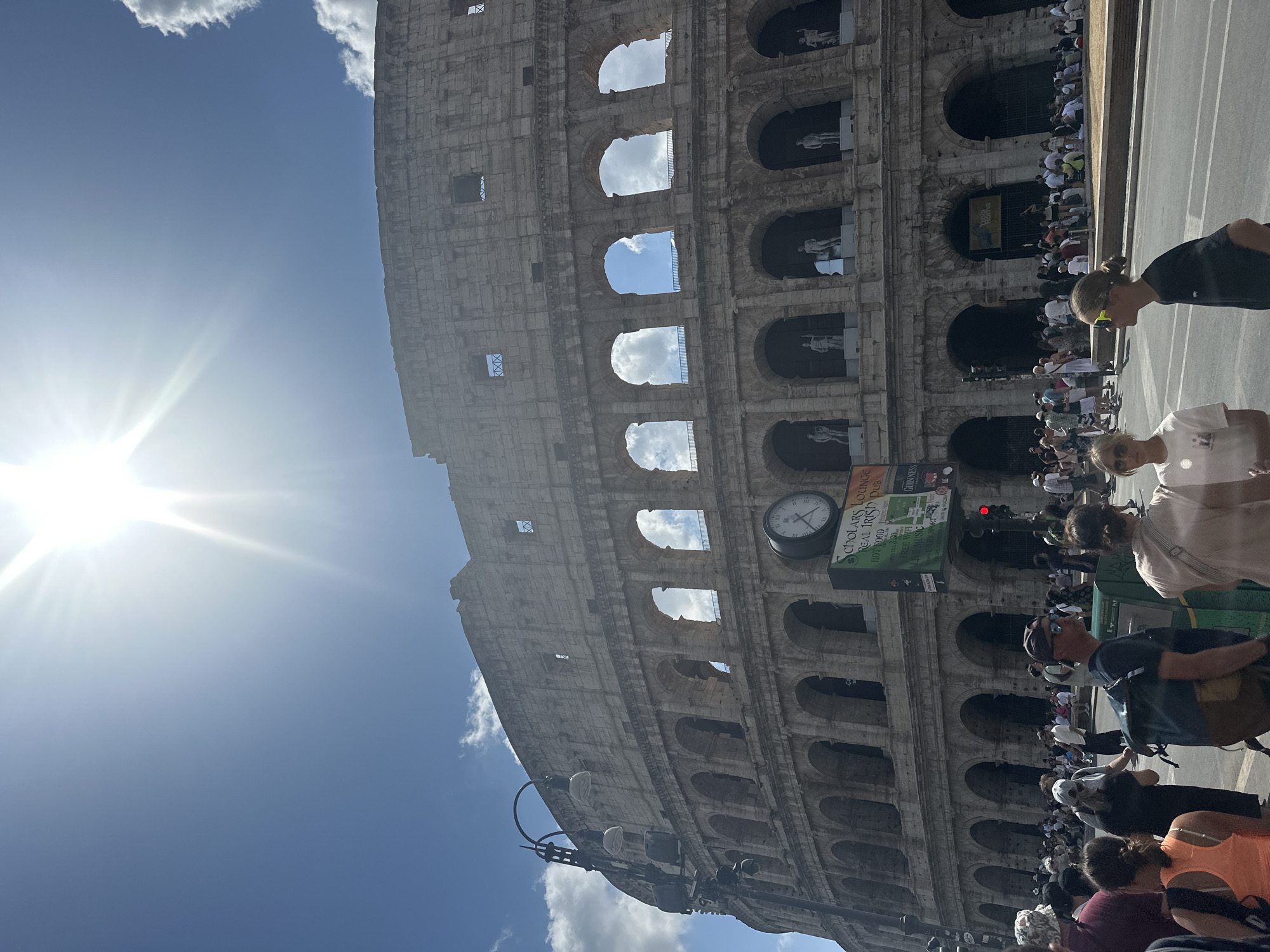 The image shows the Colosseum in Rome, Italy, under a bright, sunny sky with scattered clouds. A large crowd of tourists is visible in the foreground, walking and standing on the street in front of the ancient amphitheater. Several people are captured in motion, suggesting a busy tourist area. The Colosseum's iconic arched structure is prominent, with its weathered stone facade illuminated by the sun. A street clock and a green sign displaying "SCHOLARS LOUNGE REAL IRISH PUB" are also visible near the base of the Colosseum. The scene conveys a typical day at a major historical landmark, with many visitors observing the structure.