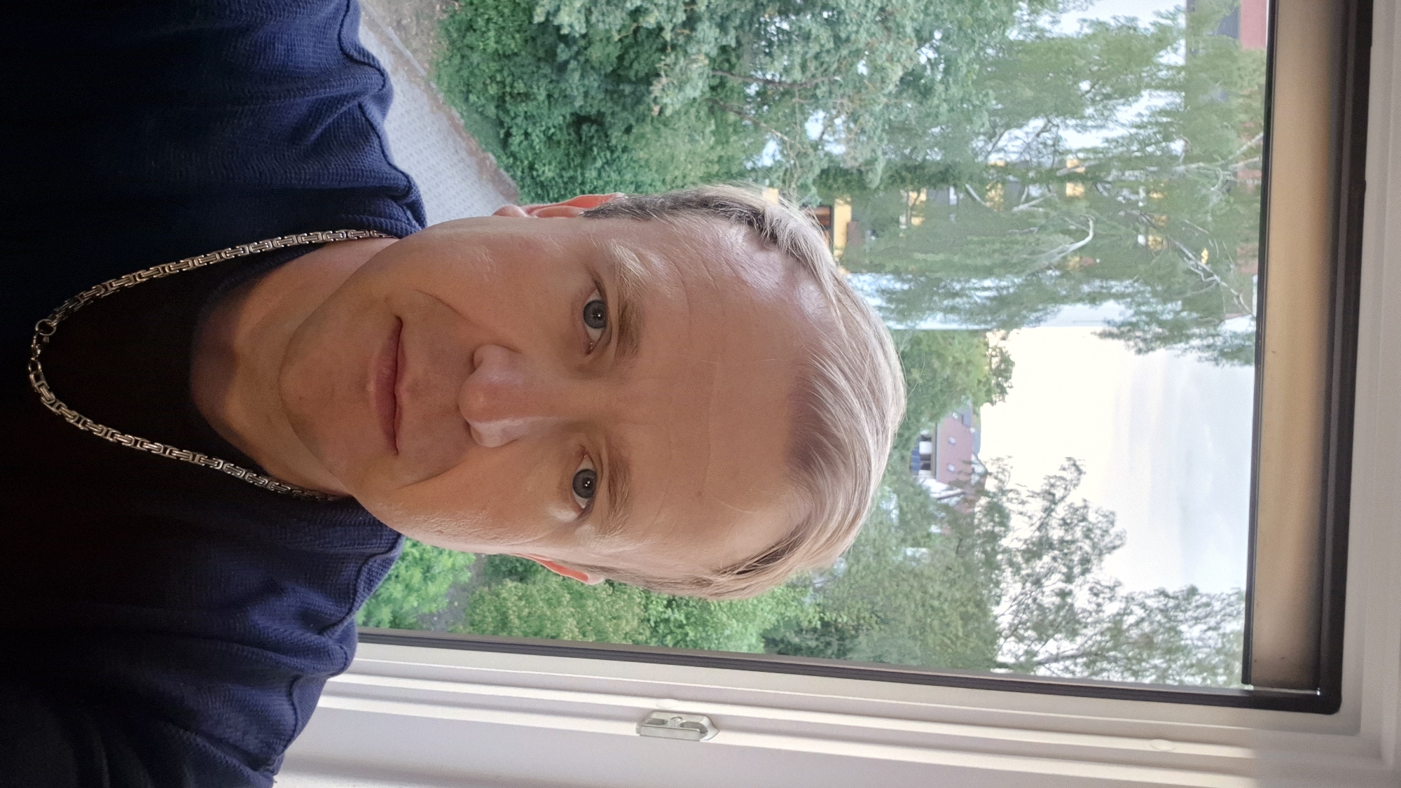 A medium close-up, eye-level indoor portrait features a light-skinned male with short, light blonde hair parted on the left, looking directly at the viewer with blue eyes. He has a neutral expression with a closed mouth. Visible facial features include a prominent nose, faint wrinkles on his forehead, and around his eyes and mouth. He wears a dark blue, possibly knitted, crew-neck shirt. A silver-colored chain with a Byzantine or King's chain link pattern is visible around his neck, extending down his chest, with the clasp visible near the bottom center of the frame.
The man is positioned in front of a window, which occupies the upper half of the background. The window frame is white on the right, and a dark brown blind is partially visible at the top. Through the window, an outdoor scene is visible under what appears to be an overcast or late afternoon sky. Lush green trees, including a weeping birch, dominate the middle ground. In the distance, parts of residential buildings with reddish-brown tiled roofs are discernible through and above the foliage. A paved pathway or road can be seen through the trees in the lower left background.