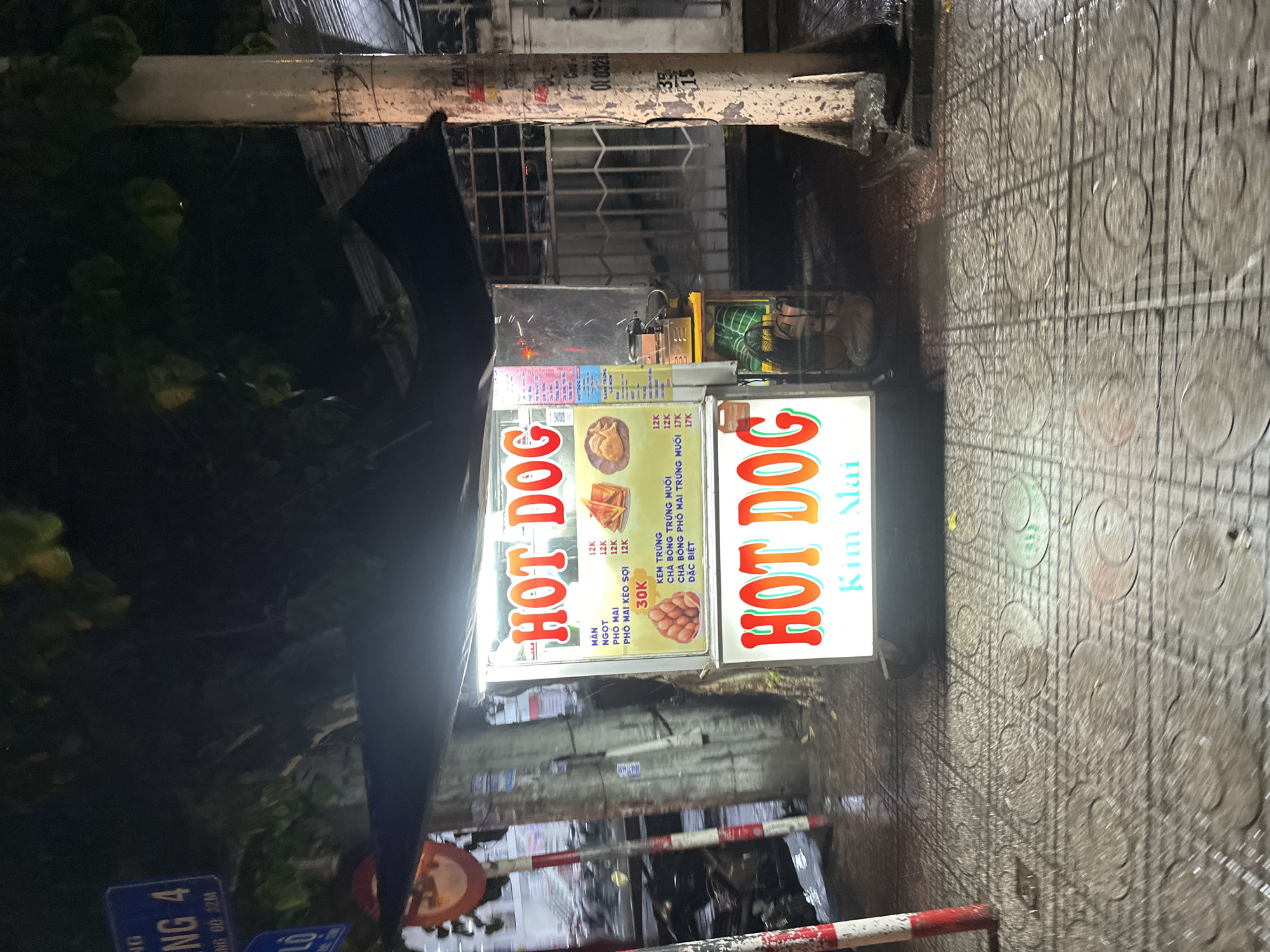 Here is a description of the image:
The image shows a street vendor selling hot dogs in Bac Nha Trang Ward, Vietnam. The vendor's cart is illuminated with signs advertising "HOT DOG" in both English and Vietnamese, with prices listed in Vietnamese Dong. Other menu items are displayed on the cart, including "PHO MAI," "PHÔ MAI KÉO SỢI," and "KEM TRÚNG". The cart is located on a sidewalk, likely on ĐÔNG HỒ Street, as indicated by the street sign visible in the lower left corner of the image. The sidewalk is wet, suggesting recent rain. The surrounding environment is dimly lit, with a building visible in the background behind a security grate. The scene is observed from a slightly elevated perspective, with some obstructions from foliage and possibly a vehicle's body.
