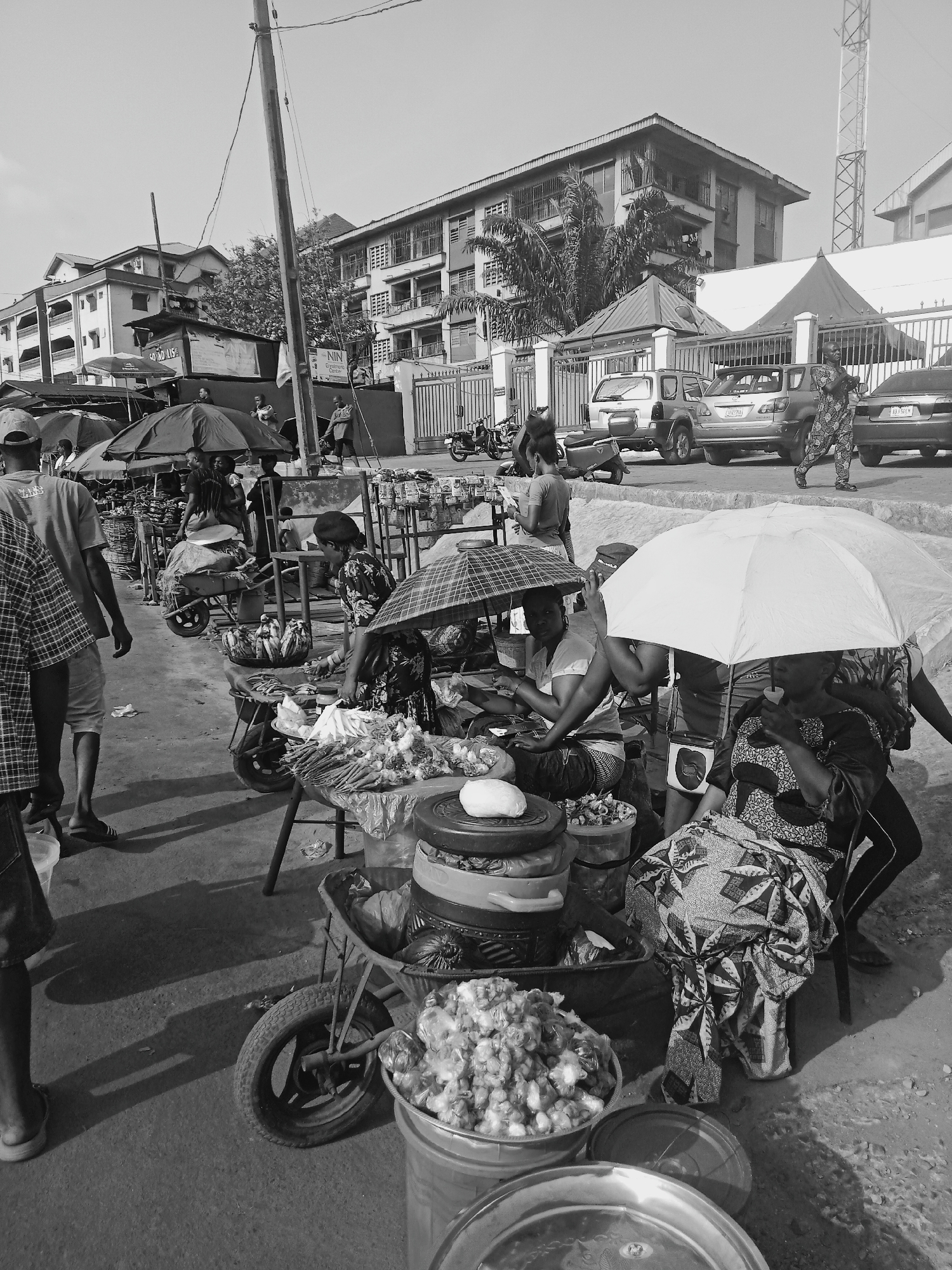 The image captures a bustling street scene in Obosi, Nigeria. Several vendors are selling goods along the street, sheltered under umbrellas. The merchandise appears to be mostly food items, including what looks like produce such as onions and other vegetables. Some of the vendors are seated, and customers are milling about, presumably shopping. A wheelbarrow laden with goods is visible in the foreground. In the background, there are buildings, cars, and pedestrians, suggesting a busy urban environment.