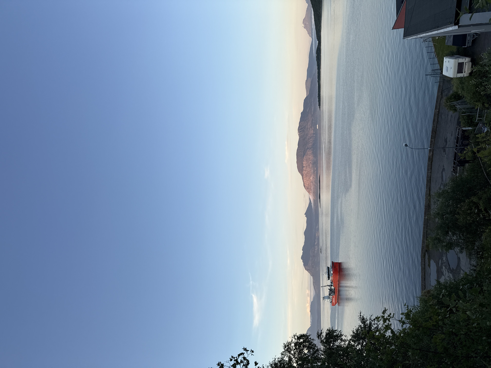 This is a wide shot taken from an elevated position, looking out over a body of water towards distant mountains under a clear blue sky. In the middle ground, a red vessel, likely a fishing or research boat, is moving from left to right across the calm water. The foreground is dominated by dark green foliage and trees on the left, and on the right, a few buildings with dark roofs and red accents, a white caravan, and a trailer are visible. The overall scene suggests a peaceful, possibly evening, atmosphere in a coastal area. The provided hints strongly indicate this is a view of the Harstad harbor in Norway, given the characteristic mountainous landscape and the presence of boats typical for the region.