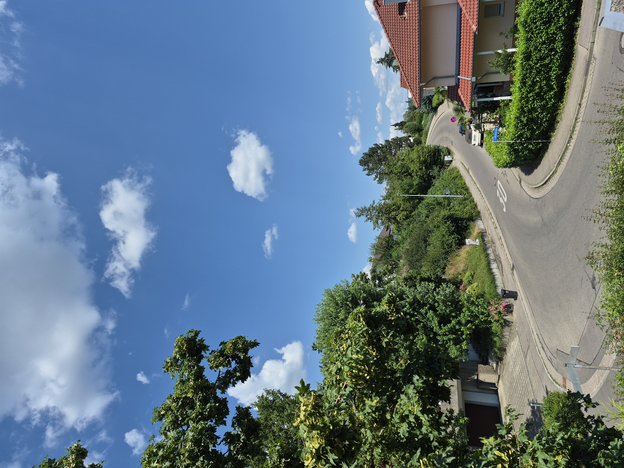 The image depicts a residential street on a sunny day. The sky is a vibrant blue with scattered white clouds. Lush green trees and hedges line the winding asphalt road. A few houses with red-tiled roofs are visible on the right side of the street. In the distance, a white van and a smaller pink object, possibly a toy or a small vehicle, are on the road. There is a blue street sign with white text and a circular sign with red and blue colors on a pole. A speed limit sign indicating "30" is visible on the road. The scene suggests a quiet suburban neighborhood in the daytime. The overall mood is peaceful and pleasant, reflecting a clear and warm day.