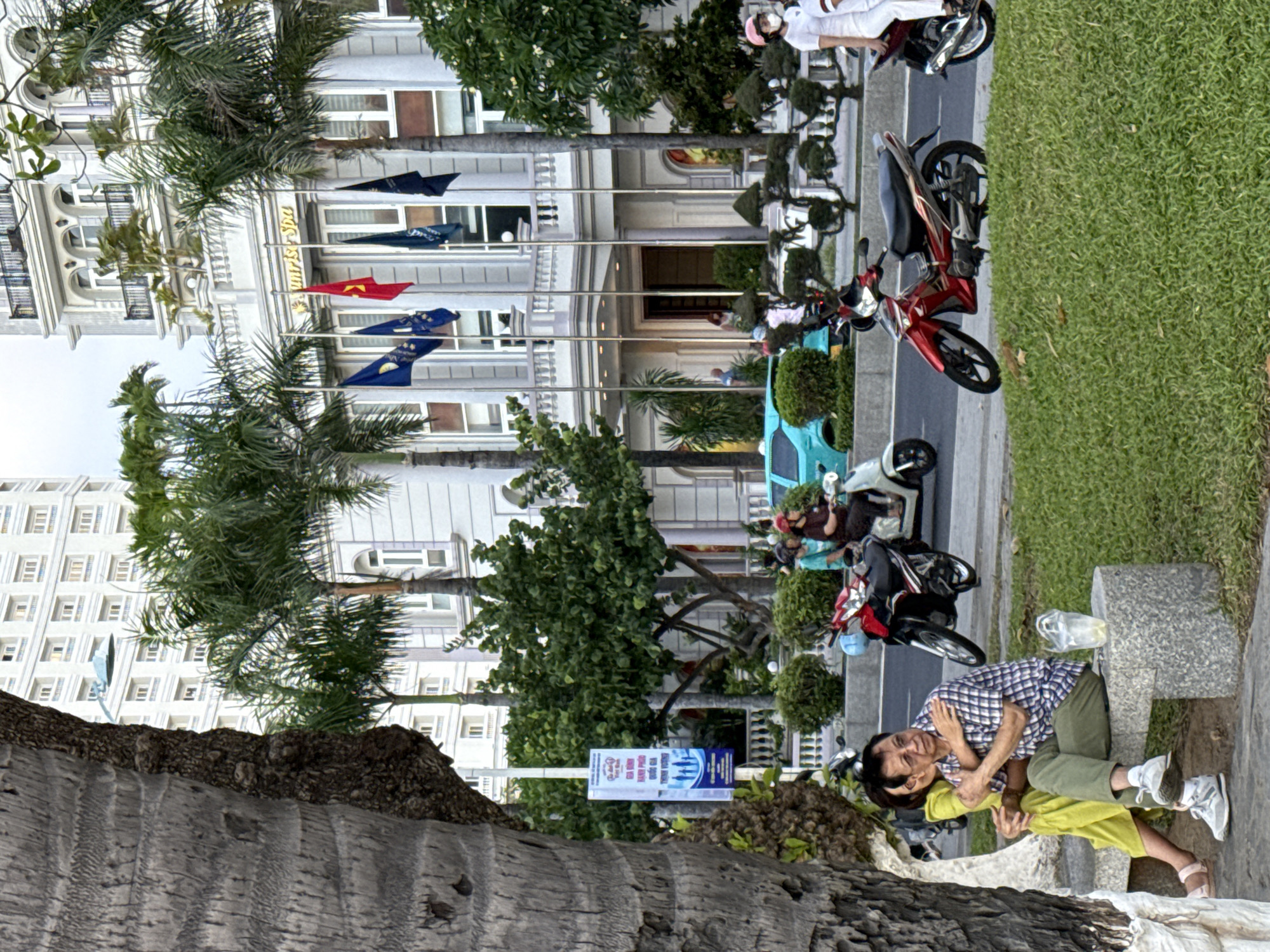 In Nha Trang, Vietnam, a couple sits on a stone bench in a park next to a street. The woman, wearing a lime-green outfit, hugs the man from behind. He wears a plaid shirt and green pants. A plastic cup sits on the bench. In the background, several motorcycles are parked and in motion on the street. Palm trees and a large, white building with flags and signage that reads "Sunrise Spa" stand behind the street. A tall tree trunk partially obscures the left side of the image.