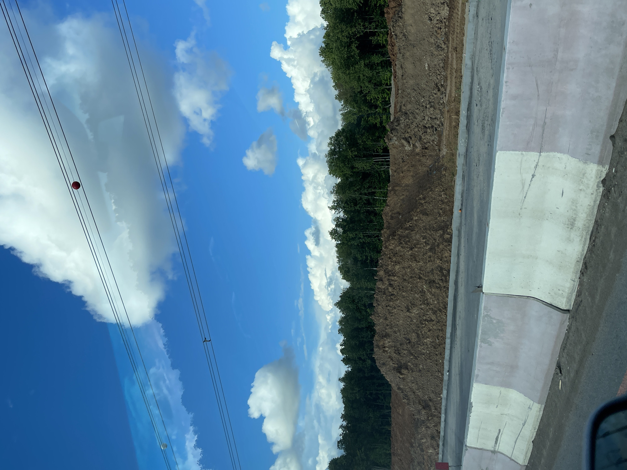 The image shows a view of the sky, clouds, trees, and what appears to be a road construction site in Novodrozhzhino, Russia. Power lines with a red marker are visible against the blue sky. White clouds dot the sky. A row of trees is adjacent to a mound of dirt. There is a gray concrete barrier along the road. The image seems to be taken from within a vehicle.