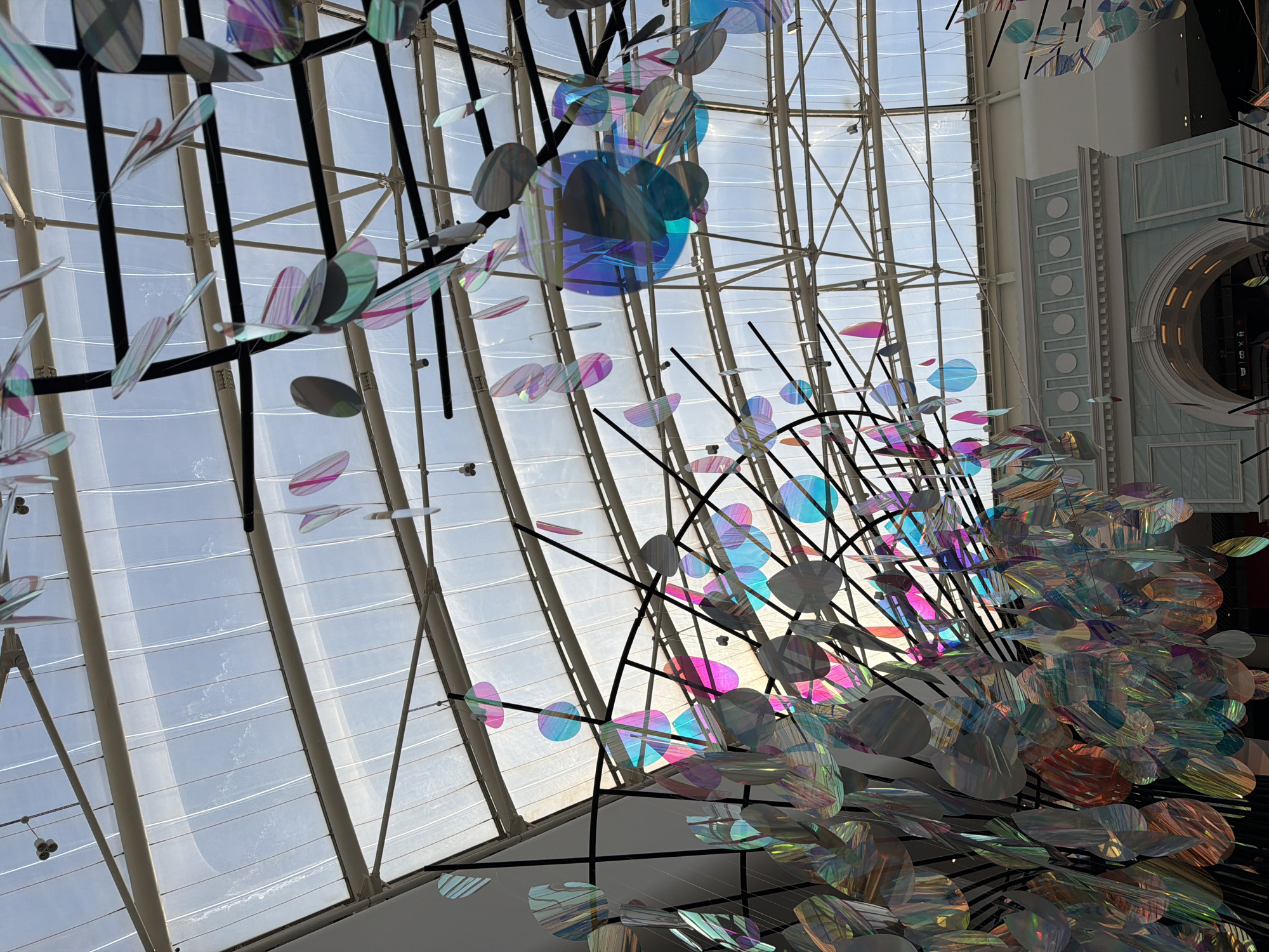 The image shows an interior view of a large, modern building, likely a shopping mall or a public atrium, with a translucent, domed roof through which daylight is filtering. Suspended from the ceiling are numerous iridescent, oval and circular decorative elements, which appear to be made of a reflective, possibly acrylic or plastic material. These elements are colorful, reflecting shades of pink, blue, green, and purple, creating a shimmering effect. The decorative structure itself is composed of black metal beams and thin wires from which the ornaments hang. Below the decorative installation, a light blue, classical-style building facade with arched openings and decorative molding is visible. The overall scene depicts a contemporary indoor space adorned with artistic light installations, suggesting a place designed for public gathering and entertainment. The location is identified as Kyiv, Ukraine.
