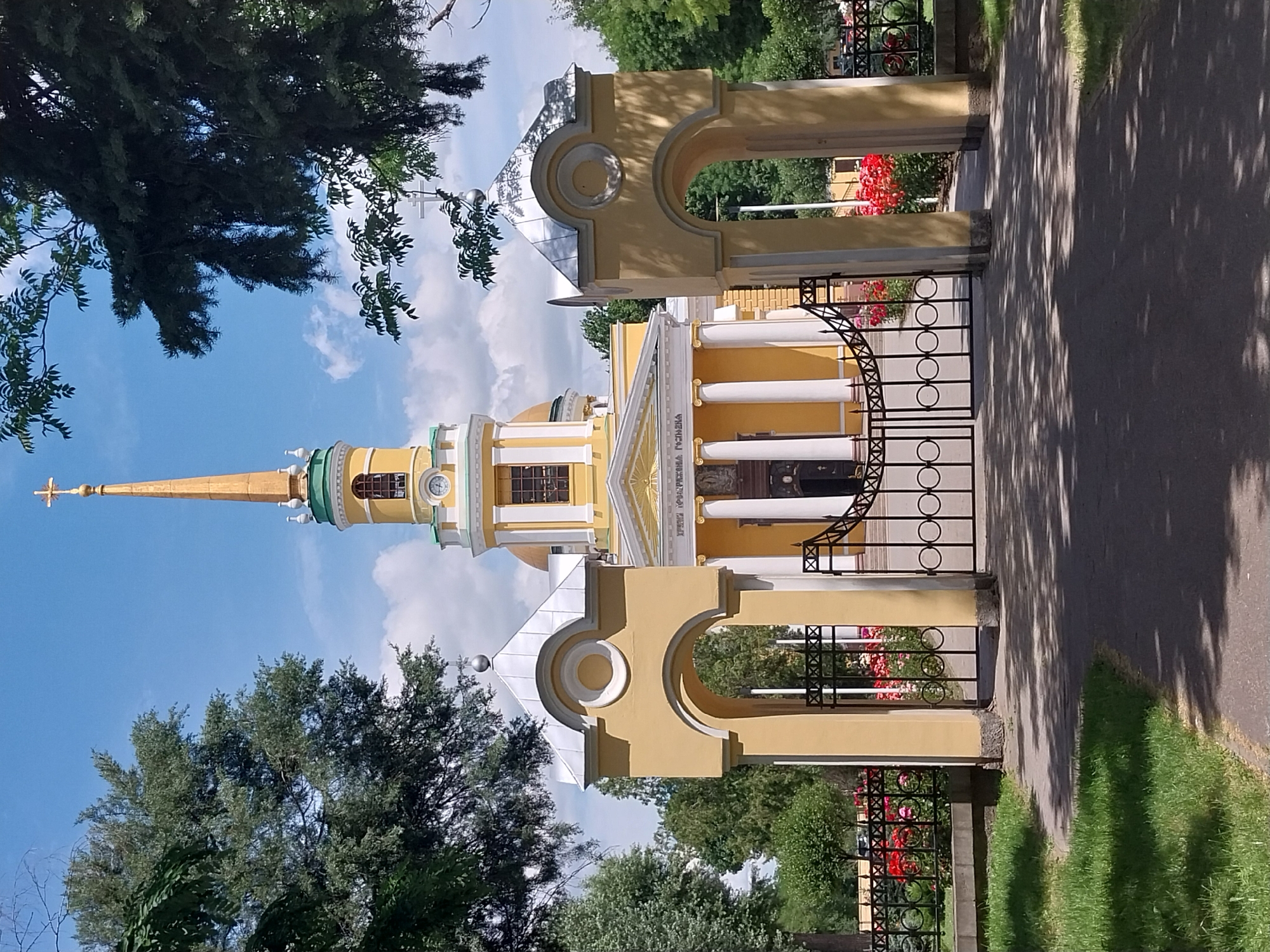 A bright yellow church with a tall, golden spire is seen under a clear blue sky with fluffy white clouds. The church features white columns and decorative architectural elements. In the foreground, a black metal gate with circular patterns separates the viewer from the church grounds, where red flowers are visible. To the right of the gate is a paved walkway partially covered in dappled shade from overhead trees. Lush green trees and grass surround the church and walkway. The image appears to be taken on a sunny day. There is text visible on the church facade, though it is difficult to read clearly. The spire is topped with a cross. The overall scene suggests a peaceful and serene setting, likely a park or religious site.