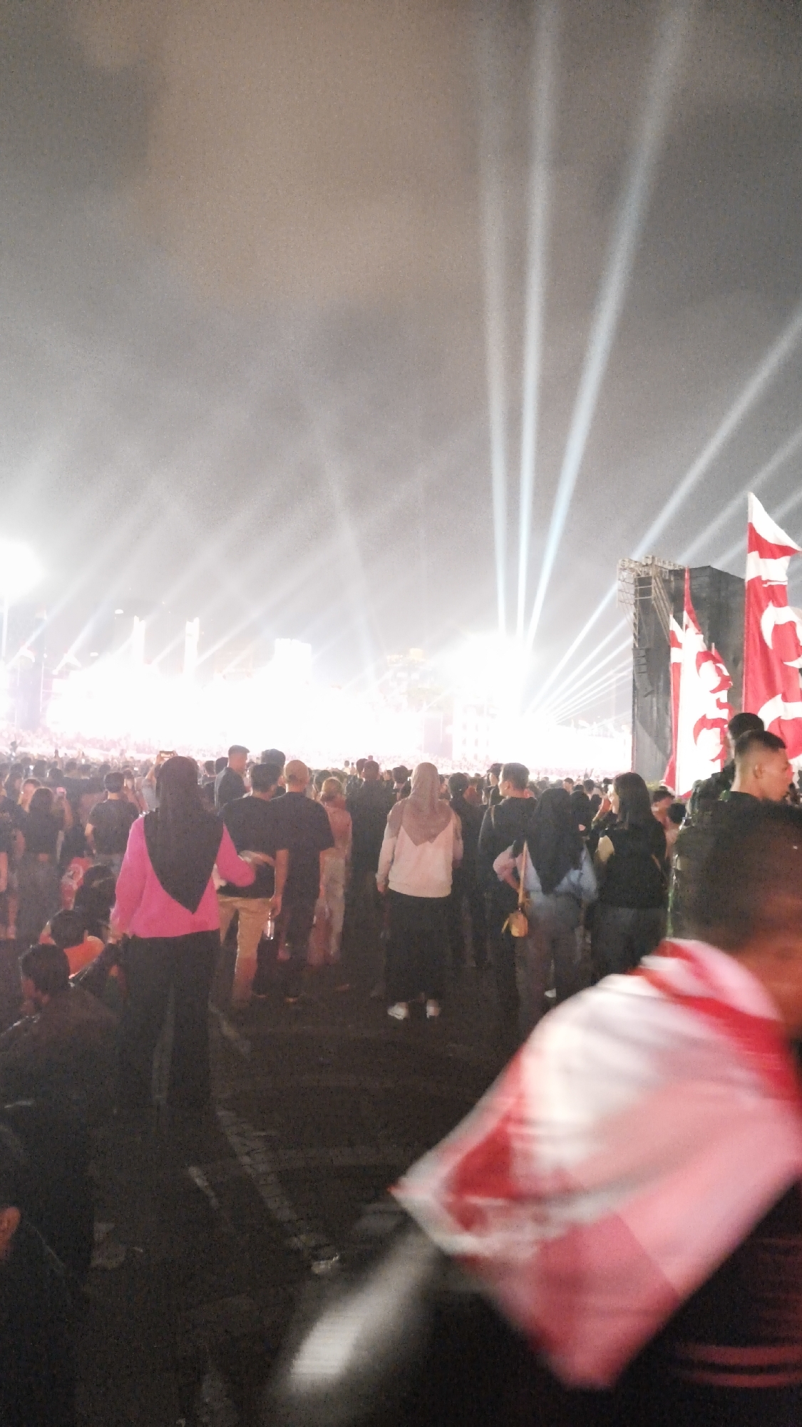 The image shows a large crowd of people gathered outdoors at night. There are bright stage lights in the background, suggesting a concert or festival. Many people are visible in the foreground and middle ground, appearing to be watching the event. Some are wearing headscarves. There are flags visible on the right side of the image. The ground appears to be dark and possibly paved. The overall atmosphere suggests a lively event.