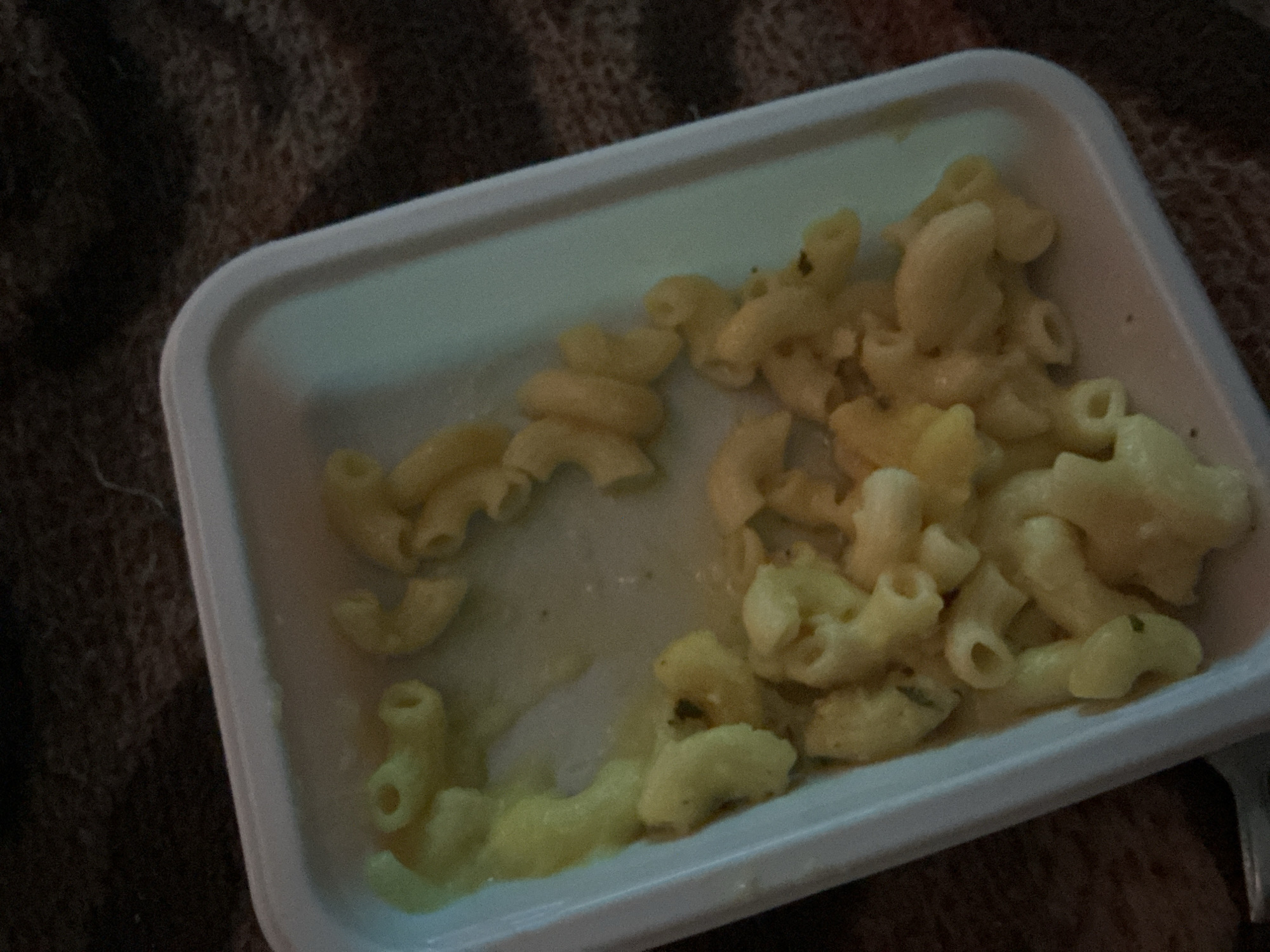 A white, rectangular plastic container holds a portion of macaroni and cheese. The macaroni pieces are curved and appear to be coated in a creamy, yellowish cheese sauce. Some pieces of dark green herb are visible mixed within the pasta. The container is resting on a dark, textured surface, possibly a blanket or rug, which has a woven pattern. The lighting in the scene is dim, casting soft shadows and highlighting the sheen of the sauce on the pasta. The image is cropped such that only the food and its immediate surroundings are visible. There is no indication of location within Merrylands, Australia, or any people or actions.