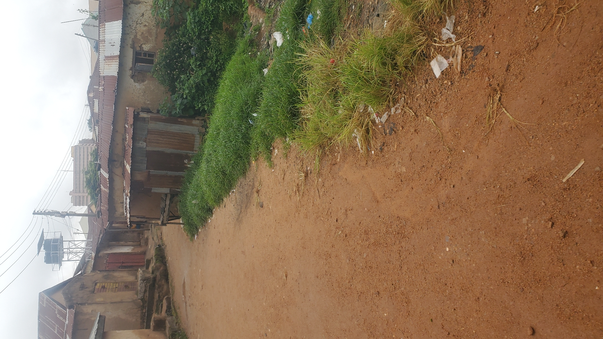 The image depicts a dusty, reddish-brown dirt road winding through a densely built-up, somewhat dilapidated urban area, likely in Jos, Nigeria. The buildings lining the road are constructed with rough, textured walls, many featuring corrugated metal roofs showing signs of rust and age. Some structures appear to be makeshift, with tin sheeting used for walls.
To the left of the road, a series of buildings are visible, some with steps leading up to entrances. One building has a red metal door. Further down the road, a tall metal structure supports two large black water tanks, indicating a public water supply system. Power lines crisscross the sky above the buildings.
On the right side of the road, a strip of overgrown green grass borders the path. This grassy area is littered with various types of debris, including white plastic bags, clear plastic fragments, and other unidentifiable trash. Behind the grass, more corrugated metal structures are visible.
In the background, beyond the immediate cluster of buildings, taller, more modern buildings with lighter facades can be seen, suggesting a contrast between older and newer parts of the city. The sky is overcast and appears a pale grey, contributing to a muted overall lighting. There are no people or animals clearly visible in the scene. The environment appears to be a working-class or informal settlement.