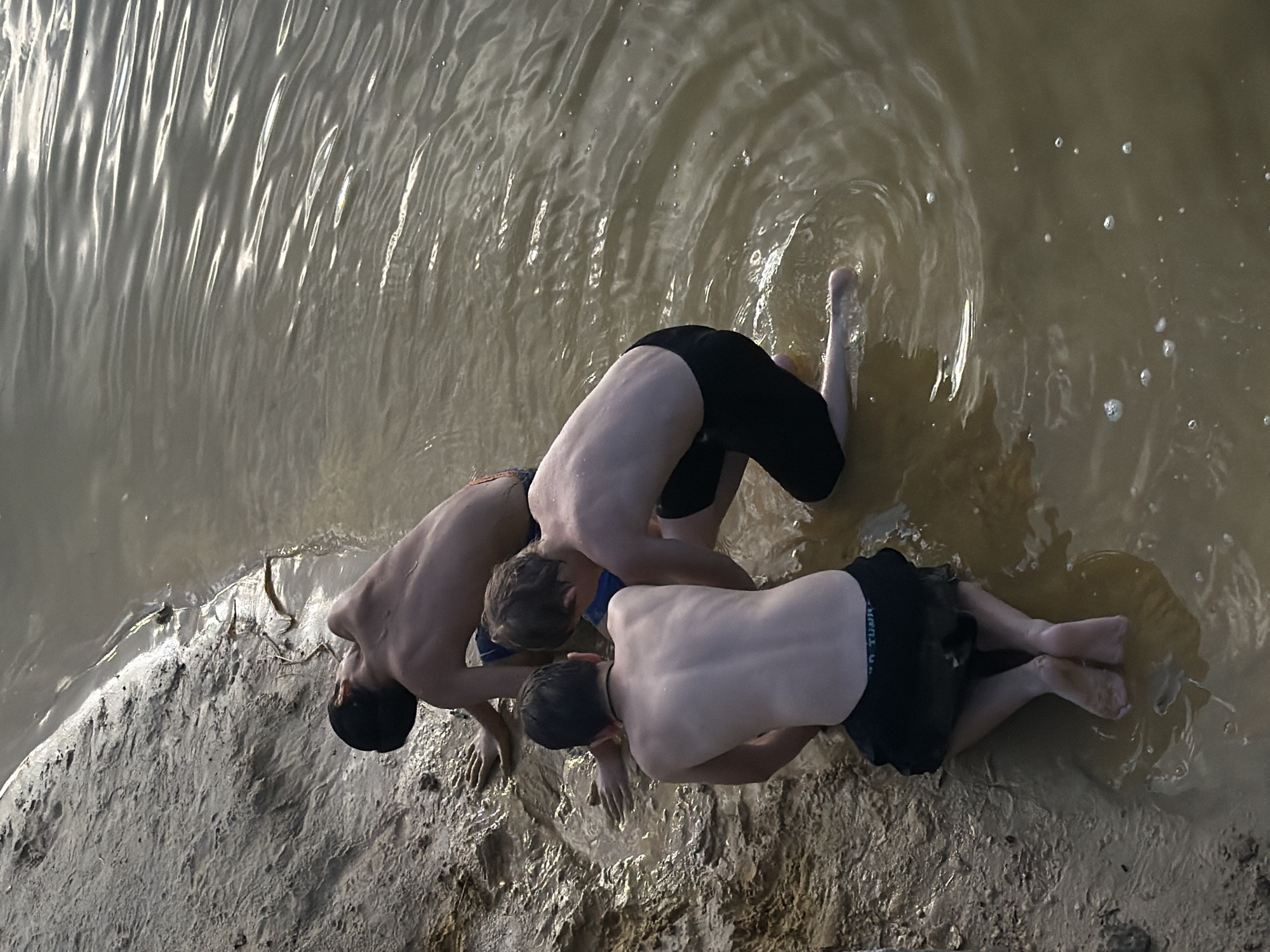 The image shows three young males in swimwear by a body of water, likely a river or lake, at the edge of a sandy embankment. The water is murky and appears shallow near the shore. The boys are interacting with the water and mud. One boy is in the foreground, kneeling with his back to the camera, his feet submerged in the muddy water. Another boy is to his left, also kneeling and reaching into the water with his hands. The third boy is further back, also in the water, with his body partially submerged and his head tilted back. The environment suggests a natural outdoor setting. The overall impression is of children playing by the water on a warm day. There are no identifiable landmarks or specific architectural elements that would pinpoint the location to Udelnaya, Russia.