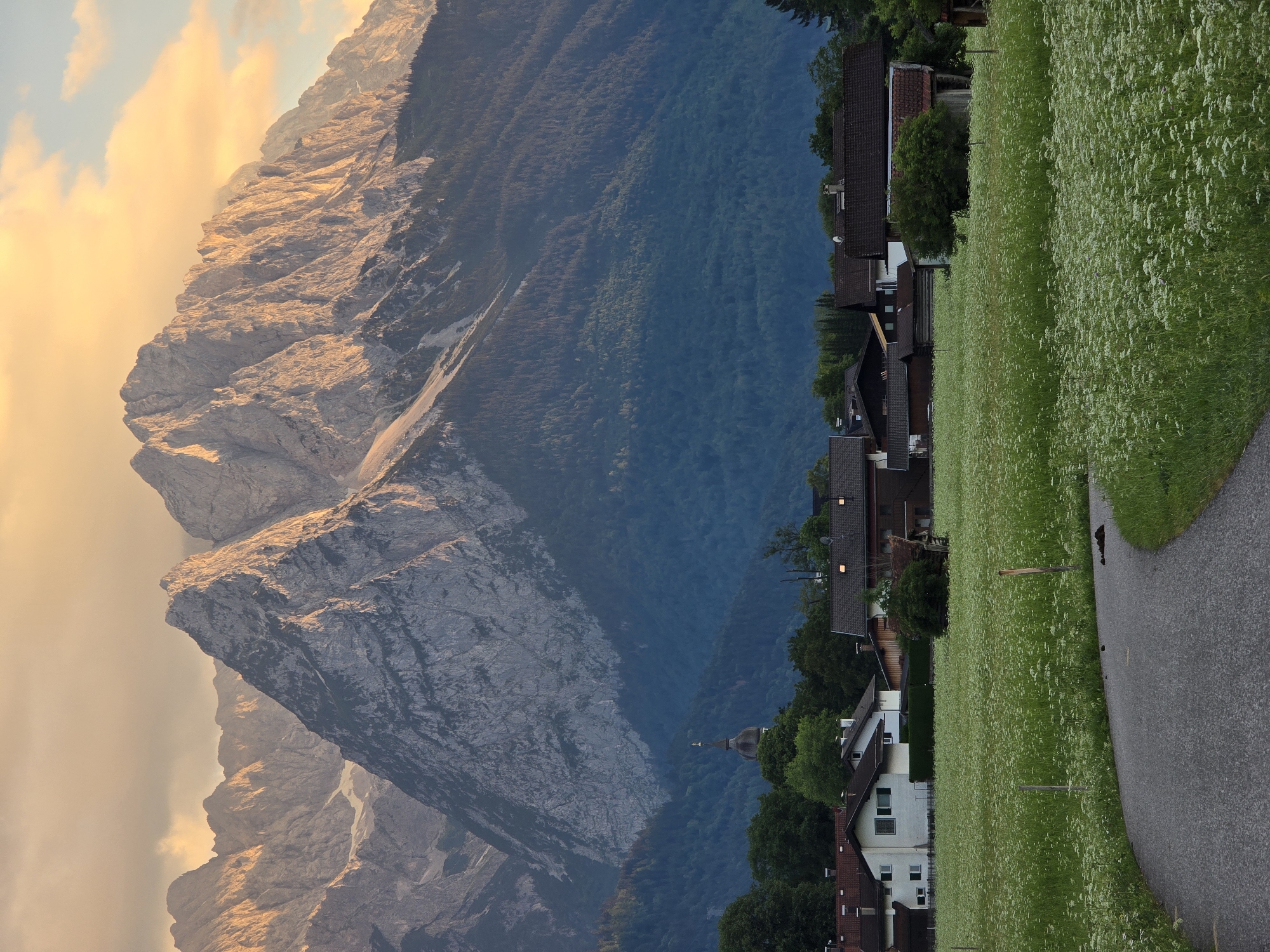 The image depicts a picturesque landscape scene in or near Garmisch-Partenkirchen, Germany, dominated by a majestic mountain range in the background. The mountains, illuminated by the warm, golden light of what appears to be either sunrise or sunset, showcase rugged peaks and slopes, some with patches of snow. In the mid-ground, a dense, dark green forest covers the lower slopes of the mountains.
Below the forest line, a small village or hamlet is nestled. Several houses with red-tiled roofs are visible, interspersed with trees and greenery. One prominent structure, a church with a distinct spire, stands out amongst the houses. The foreground features a winding asphalt road leading into a vibrant green field teeming with small white wildflowers. The field is dotted with a few wooden posts, suggesting it might be pastureland or a meadow. The overall atmosphere is serene and idyllic, characteristic of a Bavarian alpine setting. No people are visible in the image.