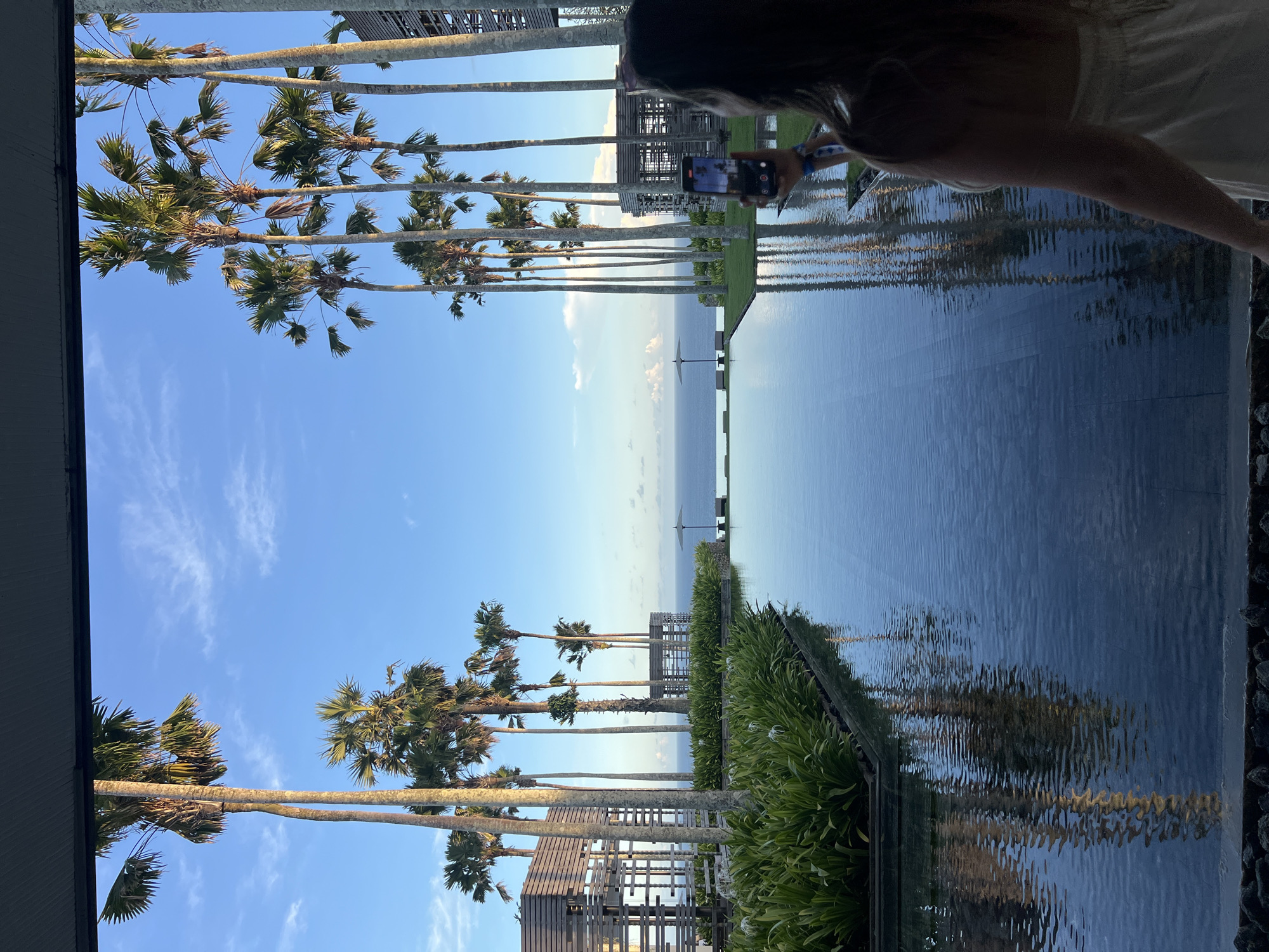 This image captures a scenic tropical resort setting in Kuta Selatan, Indonesia, showcasing a beautiful blend of natural beauty and architectural design. The photo is taken from an elevated perspective, looking out towards the ocean.
In the foreground, a person is visible from the back, positioned on the right side of the frame. They are holding a smartphone, seemingly capturing the panoramic view before them. The person has dark hair and is wearing a light-colored top, with a wristband visible on their left wrist.
Dominating the midground is a large, calm body of water, likely an infinity pool or a decorative water feature, which reflects the clear blue sky and the surrounding elements. A row of tall, slender palm trees with green fronds extends along the edge of this water body, creating a striking visual line. Beyond the palms, additional resort structures, possibly wooden pergolas or buildings, are visible.
In the background, a vast expanse of the tranquil blue ocean stretches out to meet the horizon, under a bright, mostly clear sky dotted with sparse, wispy white clouds. A white beach umbrella can be seen far in the distance, partially silhouetted against the ocean. Lush green foliage and bushes line the immediate edge of the water feature in places.
The time of day appears to be during daylight hours, with ample natural light illuminating the scene, suggesting either morning or afternoon. The weather is clear and pleasant, contributing to the serene and picturesque atmosphere. No discernible text is visible in the image. The overall impression is one of a luxurious and relaxing vacation destination.