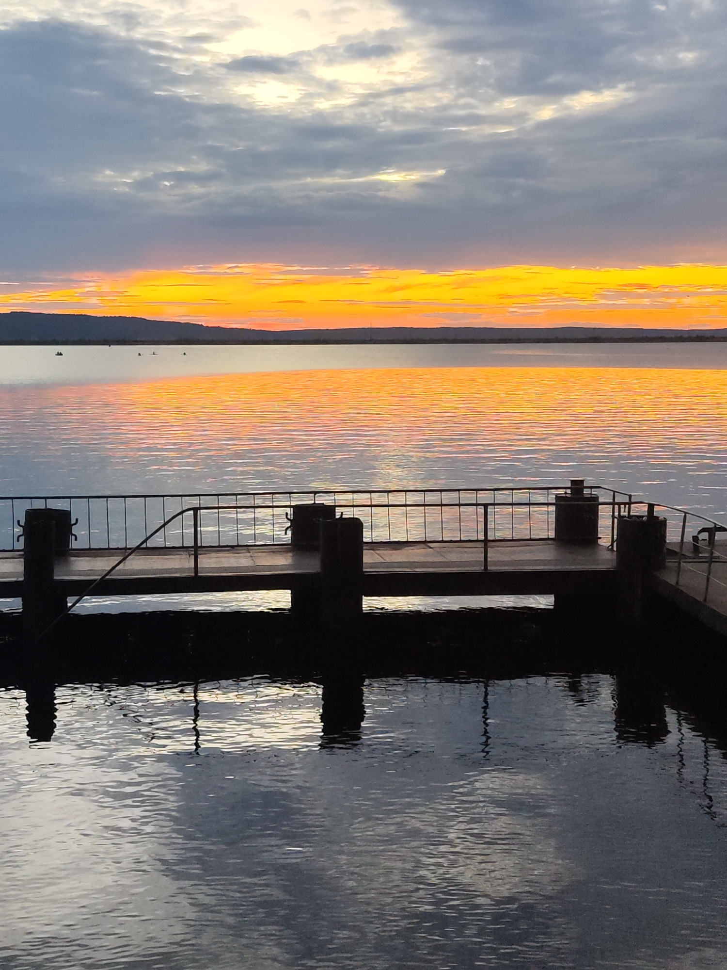 A vertical, low-angle shot captures a pier extending into a tranquil body of water at sunset. The sky displays a dramatic gradient of colors, with deep grey clouds transitioning to a vibrant band of orange and yellow just above the horizon. The distant shoreline is silhouetted against this colorful sky. The water reflects the sky's hues, with ripples creating streaks of light and shadow. The pier itself is dark and industrial, with a metal railing and thick, dark posts. The water beneath the pier is choppy, with dark reflections of the pier's structure. Several small boats are visible in the distance, scattered across the water. This scene is located in Chaikovsky, Russia, along the Kama River.