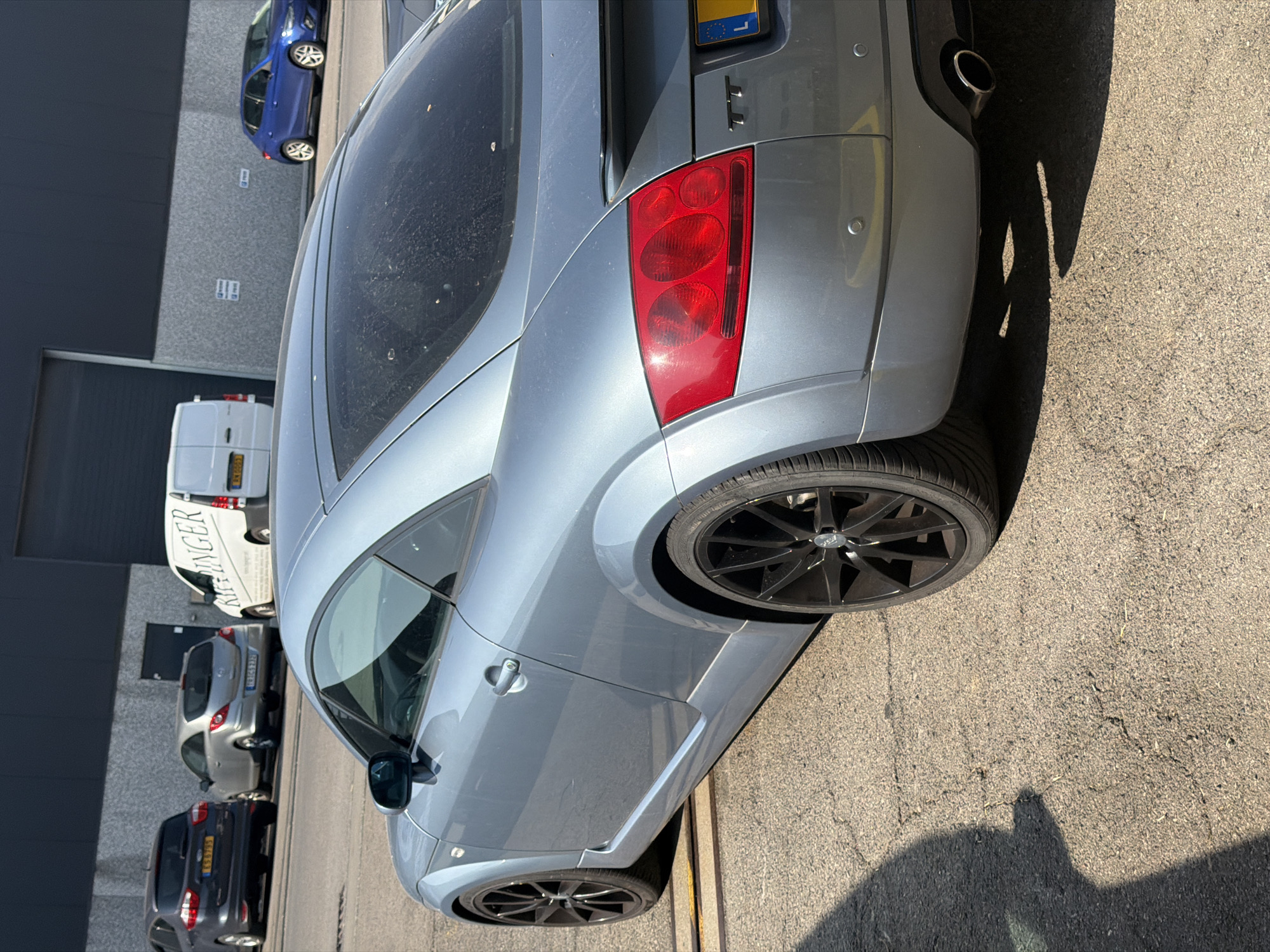 The image captures an outdoor scene in Leudelange, Luxembourg, featuring a grey Audi TT coupe parked on an asphalt surface. The car is positioned diagonally, with its rear and right side prominently displayed. The vehicle has a spoiler, distinctive taillights, and black alloy wheels. In the background, a white van with "KRINGE R" lettering is visible, along with a dark grey building with a modern facade. Other vehicles, including a blue hatchback, a silver hatchback, and a dark SUV, are parked further in the distance. The environment suggests an industrial or commercial area with a clear sky and bright sunlight casting shadows on the ground. The asphalt appears somewhat cracked and weathered.