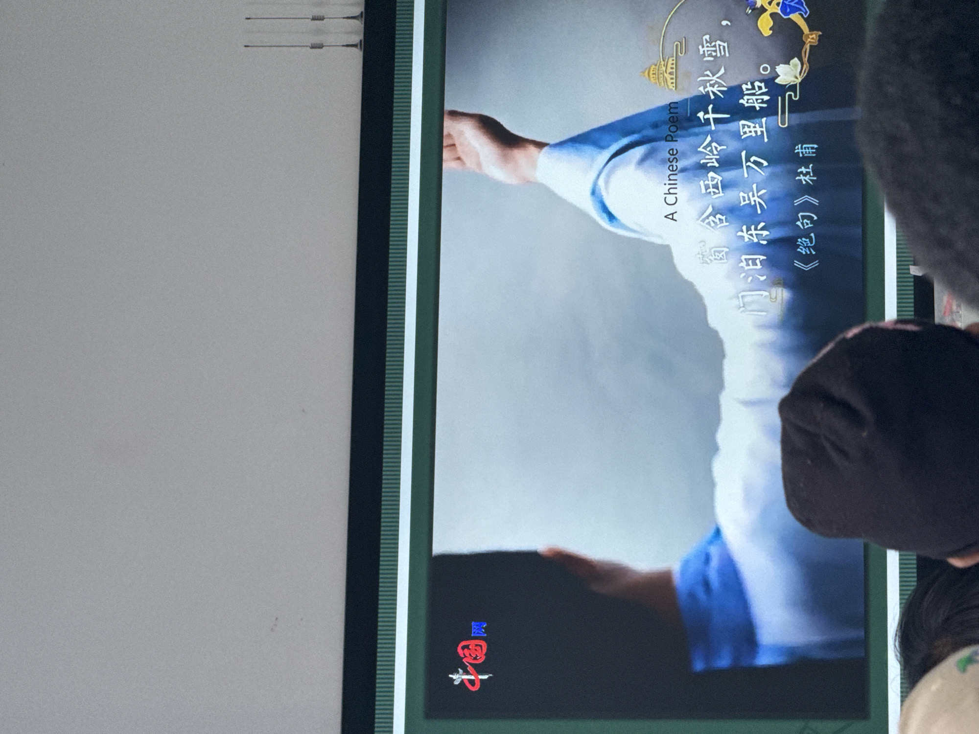 This media file captures a large screen displaying a traditional Chinese poem, likely in an indoor setting in Zhongtun, China. On the screen, a person's arm, dressed in blue and white traditional clothing, is visible, extending towards the center. Decorative elements, including a golden building and floral motifs, adorn the top right corner of the screen.
The prominent text on the screen reads "A Chinese Poem" in English. Below this, the Chinese verses "窗含西岭千秋雪," (My window frames the Western Mountains' thousand years of snow,) and "门泊东吴万里船。" (And at my door, a ship from Eastern Wu for ten thousand li.) are displayed. The poem's title and author, "《绝句》杜甫" (《Quatrain》by Du Fu), are also visible. In the bottom left corner of the screen, there is a red and blue logo, which appears to be "中国画" (Chinese Painting/Art).
To the right of the screen, the back of a person's head and shoulder, covered by dark fabric, indicates an observer viewing the content. The screen is framed by vertical dark green and light green striped borders. The overall scene suggests a cultural presentation or educational activity.