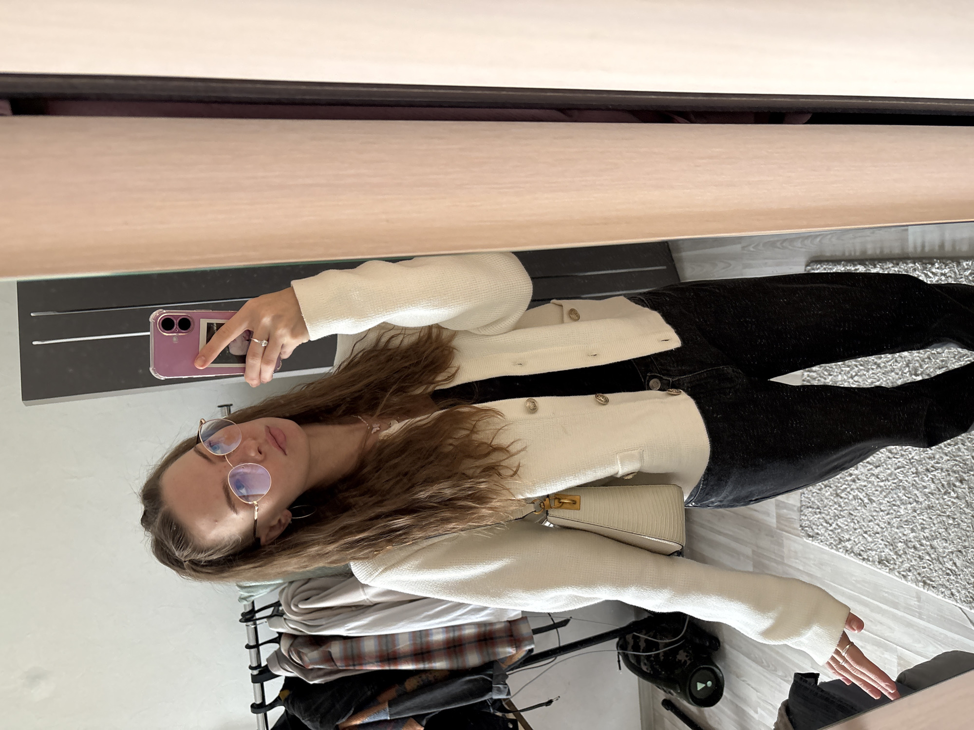 A young woman with long brown hair pulled into a half-up style and wearing round glasses is taking a selfie in a room. She is dressed in a white textured cardigan over a black top and black jeans. She is also carrying a small beige handbag with a gold clasp. In the background, there is a clothing rack with several items of clothing, a mirror, and a portion of a closet. The flooring appears to be light-colored wood with a textured grey rug. There is a floor lamp visible in the lower left corner. The scene suggests an indoor domestic environment, possibly a bedroom or dressing area. The specific location of Perm, Russia, cannot be determined from the visual information provided in the image.