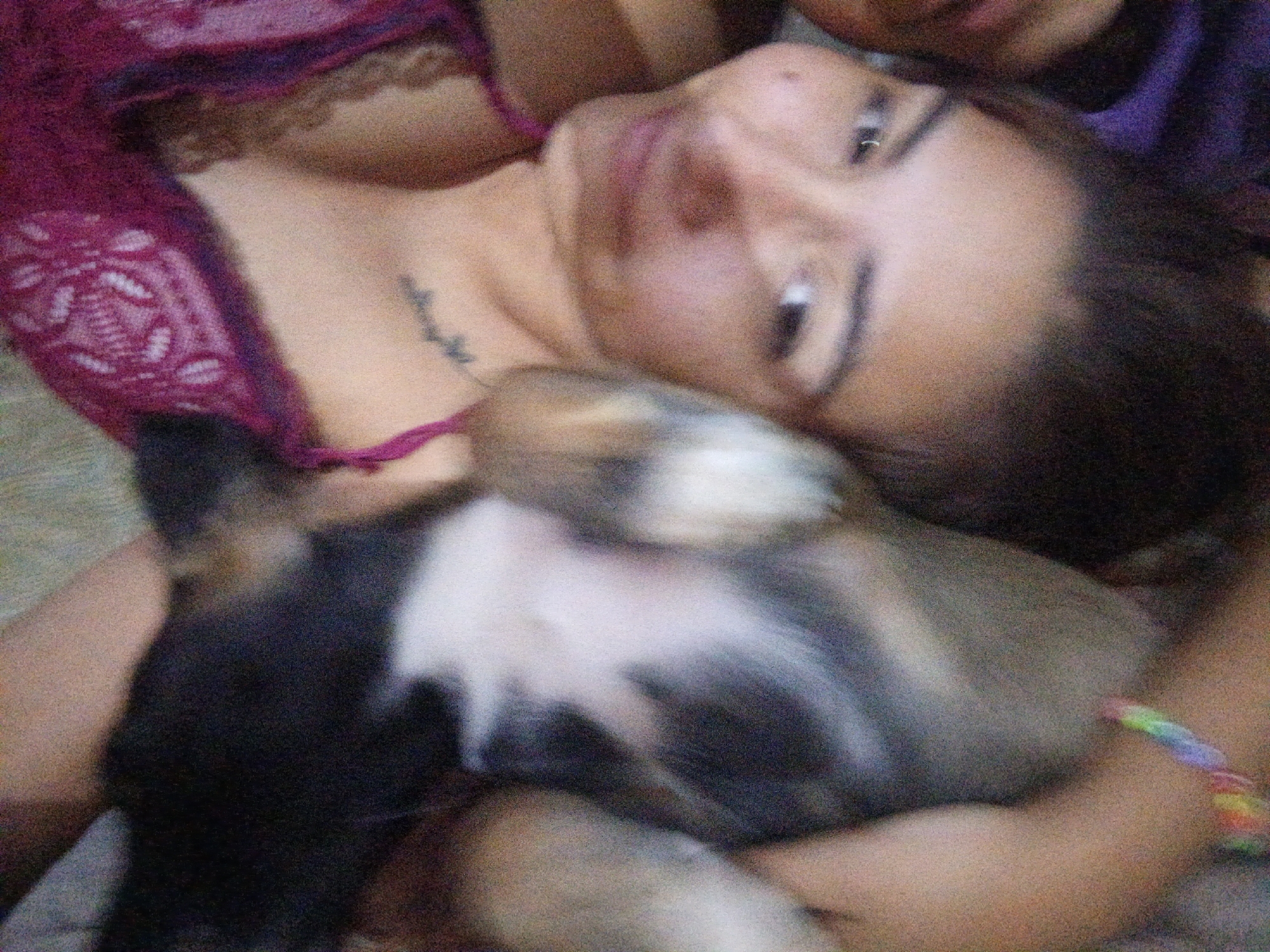 The image features a person holding a puppy. The person is lying down and is wearing a maroon lace top. They have dark hair and are looking at the camera. There is a small tattoo on the chest. The puppy is small and has a mix of brown, black, and white fur. The puppy is being held in the person's arms. A second person is partially visible in the upper right corner of the image. A bracelet is visible on the wrist. The location of the image is not discernible from the image alone. The city of Juan Griego, Venezuela cannot be confirmed.