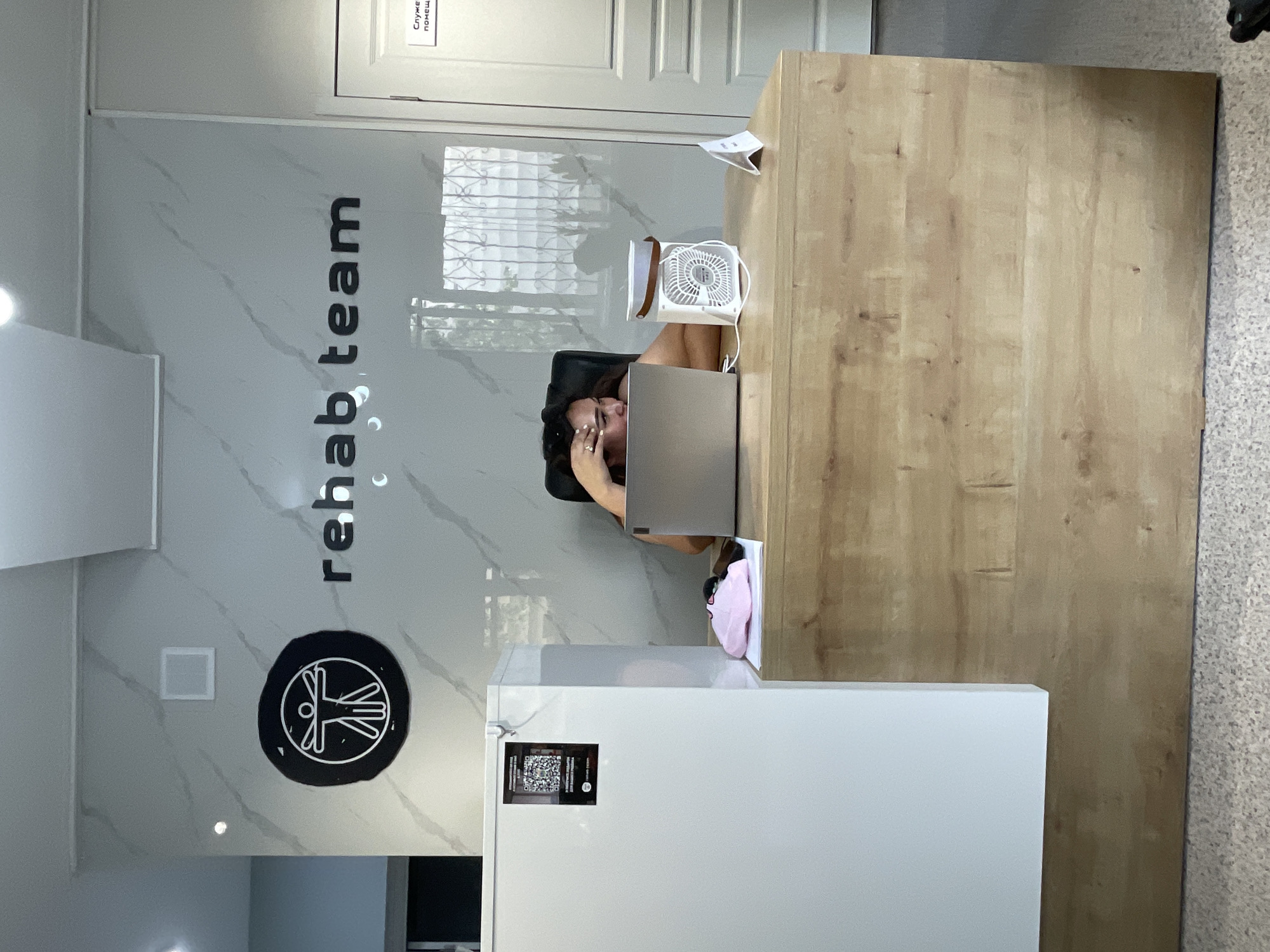 In Almaty, Kazakhstan, a woman is seated at a wooden reception desk in what appears to be a rehabilitation center. Behind her, a marble-patterned wall displays the logo of a stylized human figure within a circle and the text "rehab team" in black lettering. The woman is looking down at a laptop, her hand resting on her forehead as if she is stressed or tired. A small white fan sits on the desk next to her. To her left, a large white refrigerator is positioned by the desk, with a QR code sticker attached to its front. A pink cap and a black object, possibly a phone, are also on the desk. A door with a sign that reads "Служебное помещение" (Service room) is visible to the right of the reception area. The overall environment suggests a professional setting, possibly an office or clinic reception.