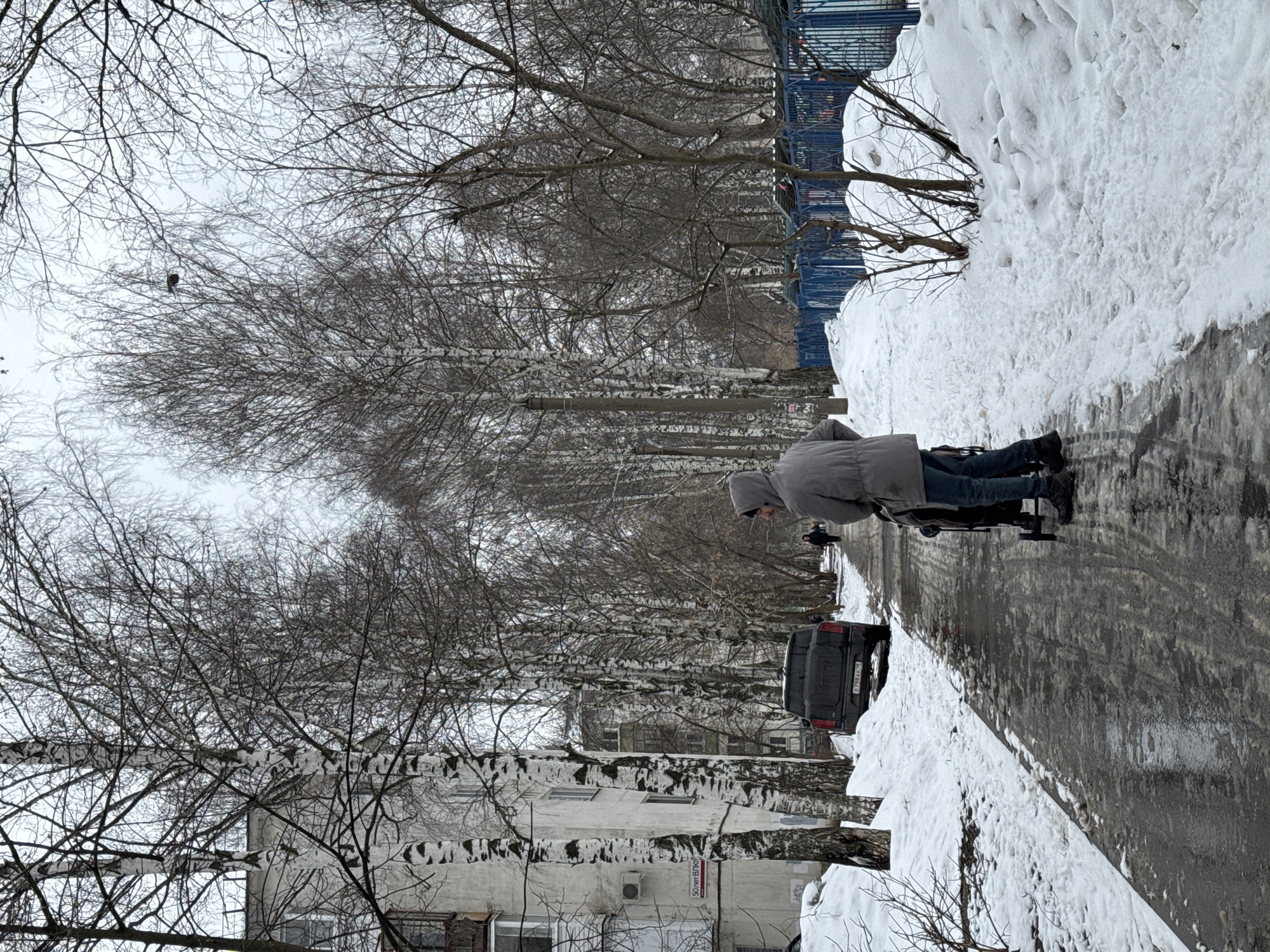 The image depicts a winter scene in what appears to be a residential area of Izhevsk, Russia. A person wearing a gray jacket and jeans is walking with a stroller on a narrow, wet sidewalk partially covered with snow. A dark-colored car is parked on the side of the road. On the left, there is a multi-story building with bare trees in front. The trees are lined along the street, creating a corridor-like effect. On the right, there is a large snowdrift. A blue fence is visible in the background. The sky is overcast.