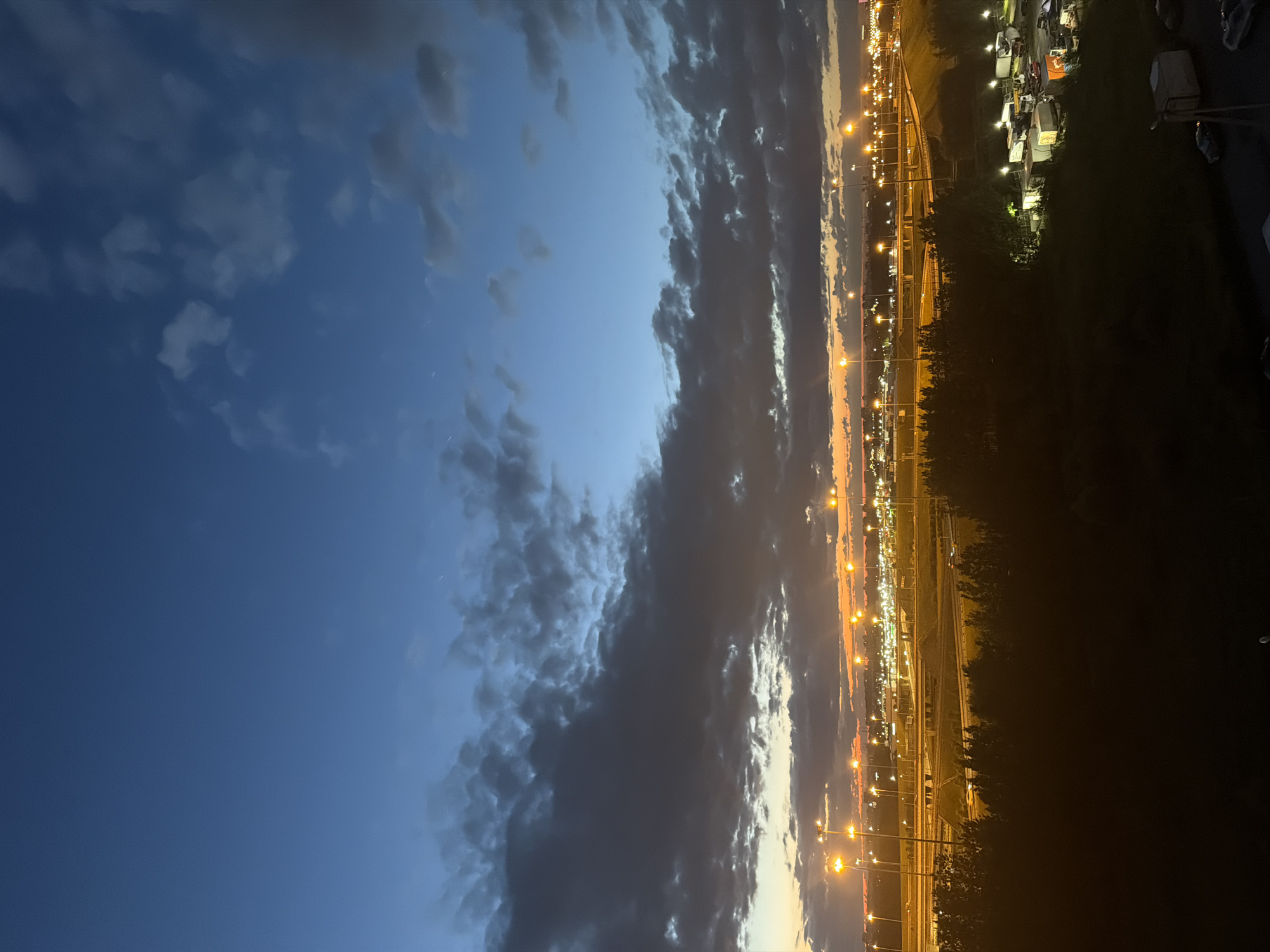 An aerial, vertical shot captures a twilight scene of a city with a vast expanse of blue sky and scattered clouds. In the foreground, a dark silhouette of trees frames the bottom of the image. Beyond the trees, a dimly lit cityscape is visible, characterized by numerous streetlights illuminating highways and industrial or commercial buildings. Several vehicles, including trucks and what appear to be trailers, are parked in a lot to the right of the frame. The horizon glows with the warm colors of sunset peeking through the clouds. The overall mood is serene and tranquil, with the artificial lights of the city beginning to counter the fading daylight.
