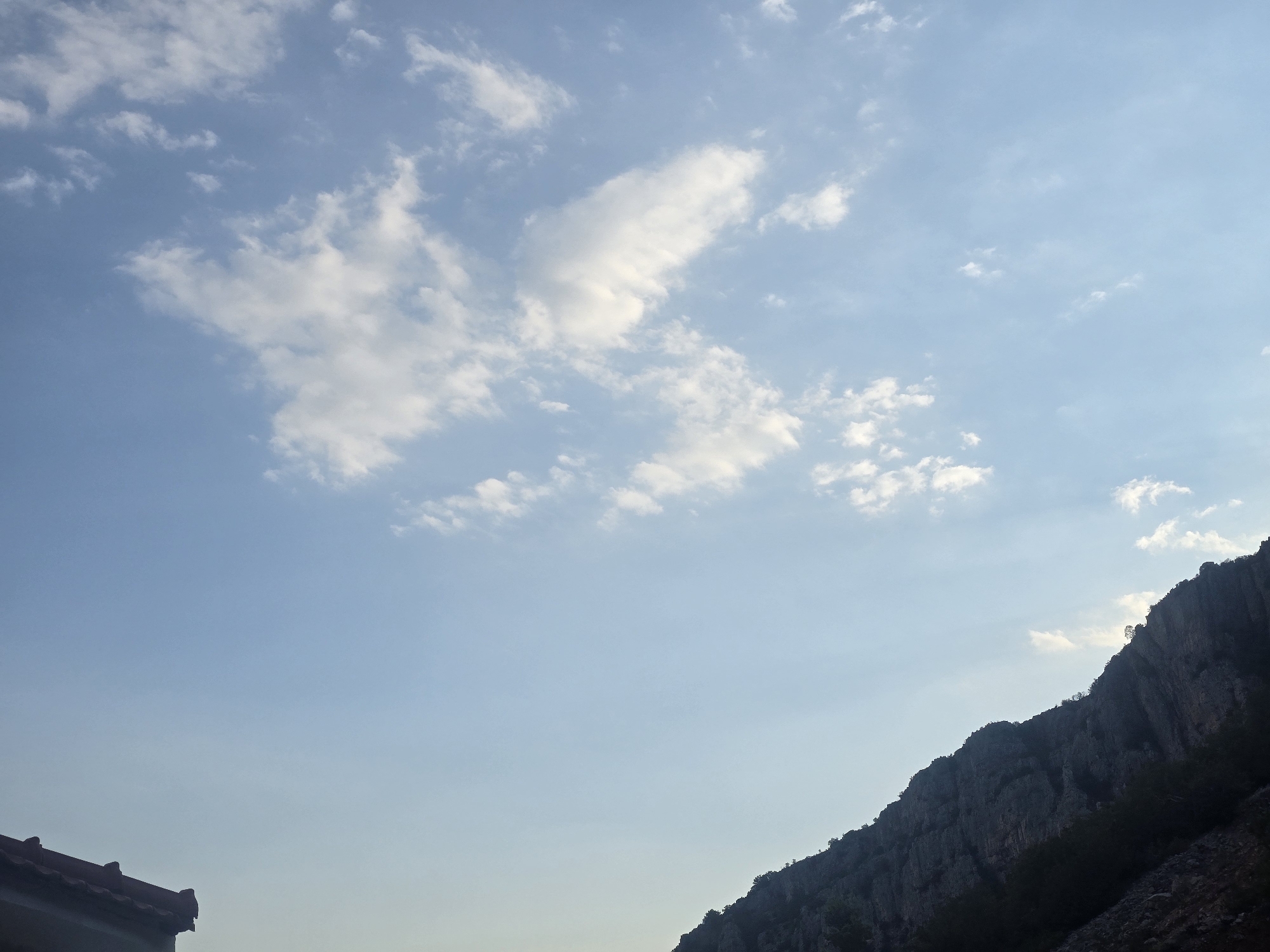 The image captures a section of a rocky mountain range, characterized by its steep, rugged slopes. The mountain appears to be covered in sparse vegetation, with some trees visible along its upper ridges. The sky above is a clear blue, with scattered white clouds, suggesting fair weather. In the lower left corner of the image, a portion of a building's roof is visible, with red tiles and dark eaves. The overall scene depicts a natural landscape adjacent to a built structure, likely situated on the outskirts of a city. While the specific location in Podgorica, Montenegro, is not explicitly identifiable from the visual elements, the scene is consistent with mountainous terrain found in the vicinity of the city. There are no people or discernible actions taking place in the image.