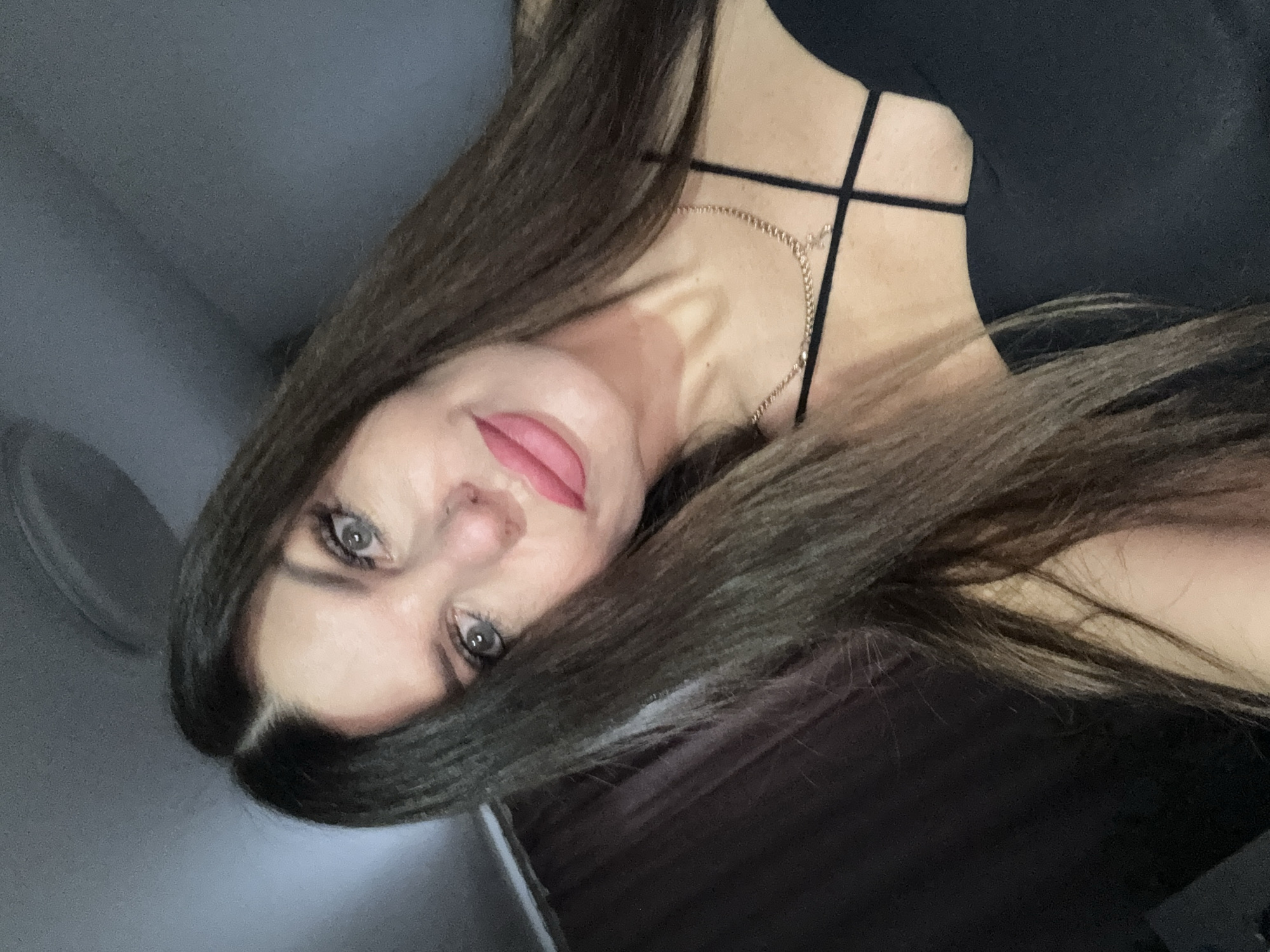This is a selfie of a Caucasian woman with long, dark brown hair and fair skin. She is wearing a black top with thin straps and a delicate gold necklace with a small cross pendant. Her makeup includes prominent false eyelashes and glossy pink lipstick. Her eyes are wide open and looking towards the camera. The background is dimly lit and appears to be indoors, with a ceiling fan visible in the upper portion of the image and a dark curtain on the left. There is no discernible information in the image that places it in the city of Chaikovsky, Russia.