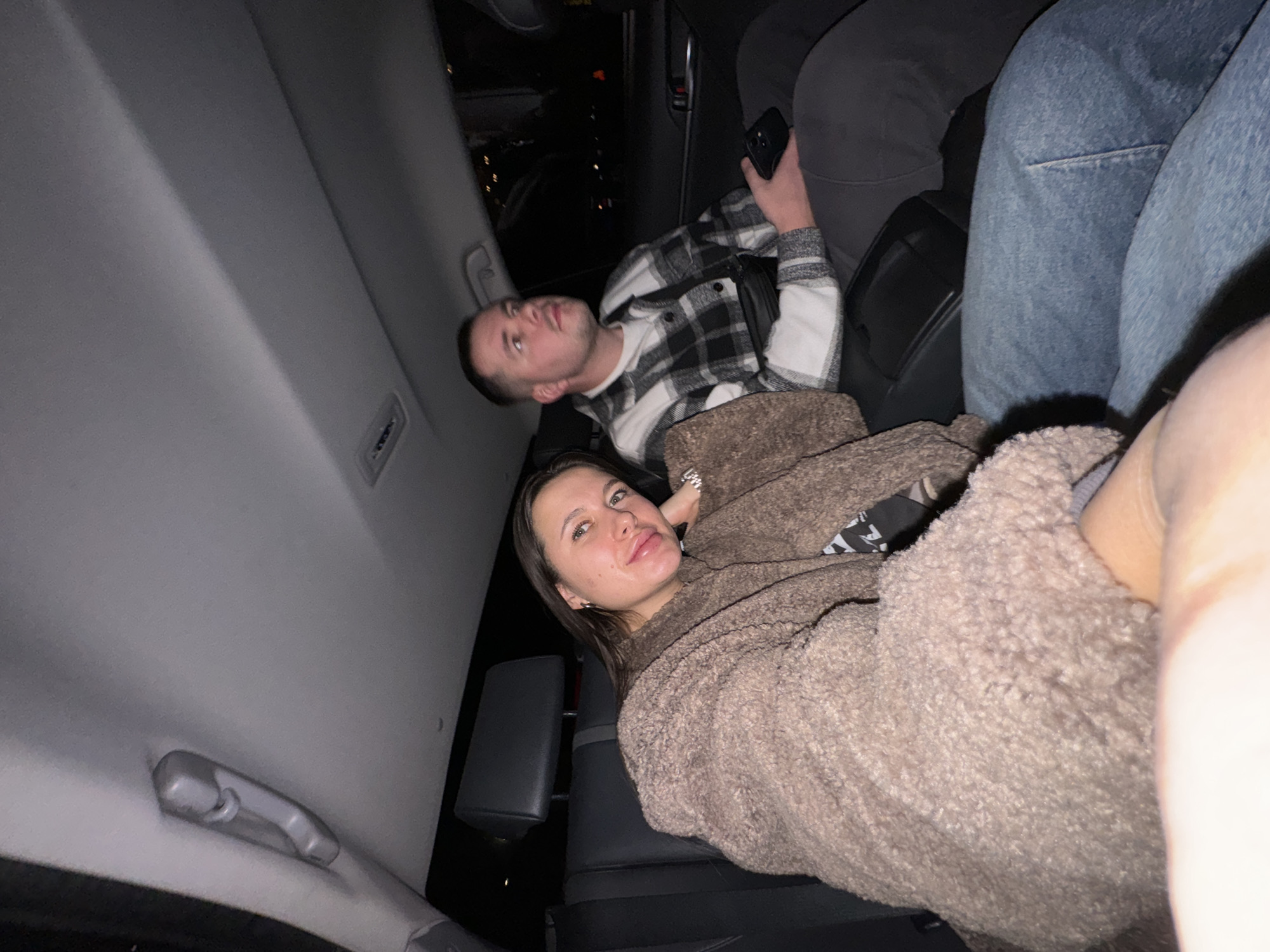 Two individuals are observed in the rear passenger compartment of a vehicle at night in Lesnoi Gorodok, Russia. A female, approximately 20-30 years old, is seated on the left, wearing a brown textured coat over a dark garment and blue jeans. Her head is tilted, resting on her right hand, and she looks directly at the camera with a slight smile. To her right, a male, also approximately 20-30 years old, is seated, wearing a black and white plaid shirt over a white undershirt, dark sweatpants, and a black fanny pack. He holds a black smartphone in his left hand and gazes forward and slightly upward, away from the camera. The car interior features dark upholstered seats with headrests, a light grey ceiling, and an overhead grab handle. The external environment visible through the windows is dark, indicating nighttime conditions.
