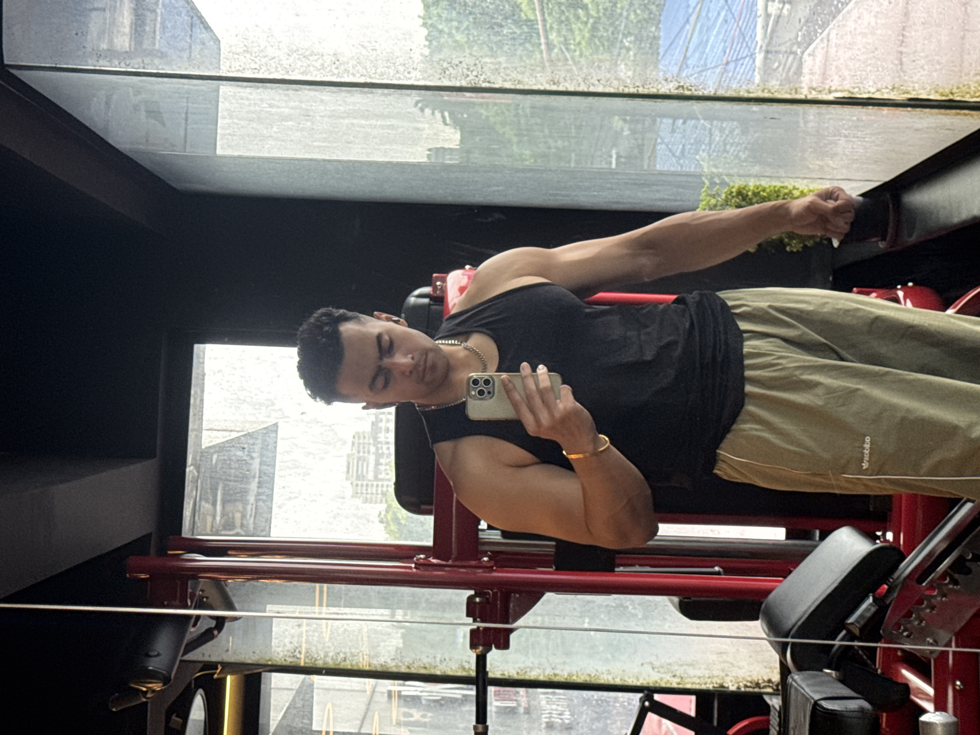A man is taking a mirror selfie in a gym, likely in Kolkata, India. He is wearing a black tank top and light-colored athletic pants. He has a gold chain around his neck and a gold bracelet on his left wrist. He is holding a smartphone in his right hand, capturing the reflection. The gym appears to have modern equipment, with a red weight machine visible in the background to the left. Large windows line the right side of the gym, offering a view of an urban environment, possibly with buildings and a blue roof in the distance. The windows appear to be wet, suggesting it might be raining outside, which is a common occurrence in Kolkata during certain seasons. The overall impression is of a fitness enthusiast documenting their workout in an urban gym setting.