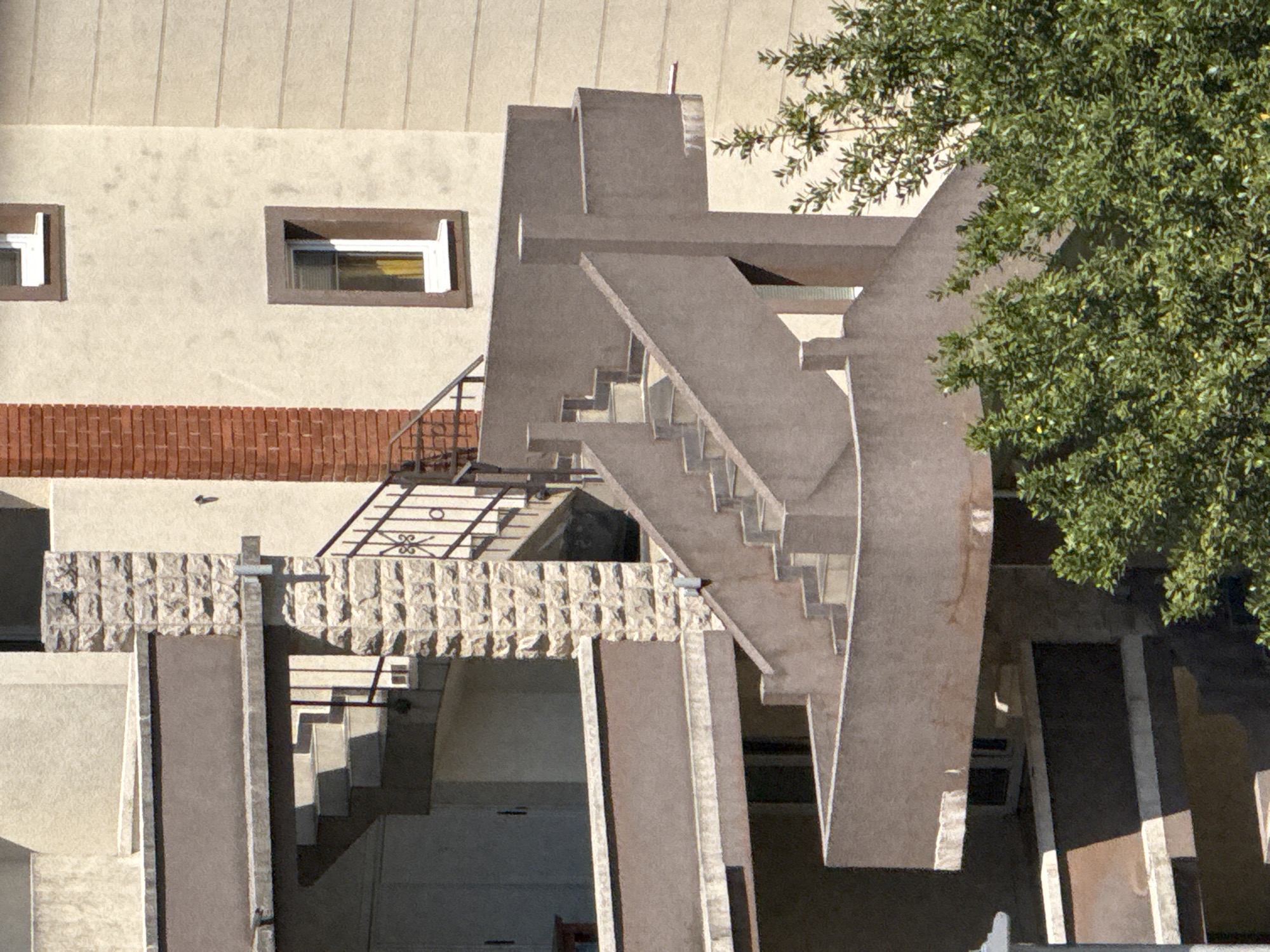 The image shows a multi-story building with an outdoor staircase. The building has a cream-colored facade with some sections of textured, light-colored stone and a vertical column of reddish-brown brick. There are several windows visible, some with brown frames and white sills. The staircase is constructed of concrete and appears to be a complex, multi-level structure with landings and railings. In the foreground, there is a dense green tree with many small leaves. The scene is likely an exterior shot of a residential or commercial building in a sunny environment, potentially in Budva, Montenegro, given the architectural style and vegetation. There are no people or animals visible in the image.