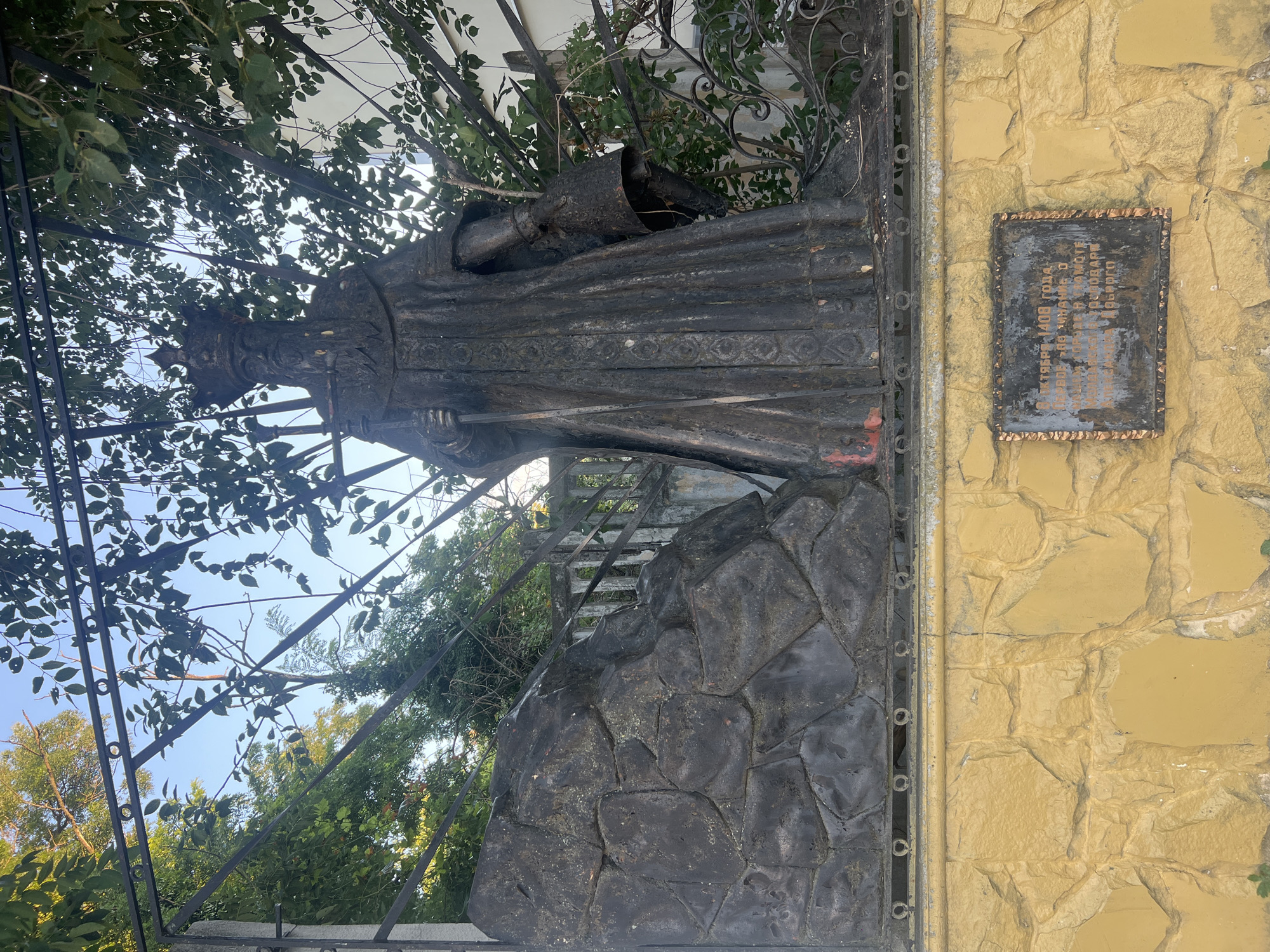The image shows a bronze statue of a man wearing a crown and a long robe, holding a staff. The statue is located in Bender, Moldova. The statue is positioned next to a large rock sculpture and is enclosed by a metal fence. In the background, there are trees and a blue sky. A plaque with text in Cyrillic script is mounted on a yellow, stone-like wall below the statue. The statue appears weathered, with some reddish-brown marks on the robe. The overall scene suggests an outdoor monument or historical site.