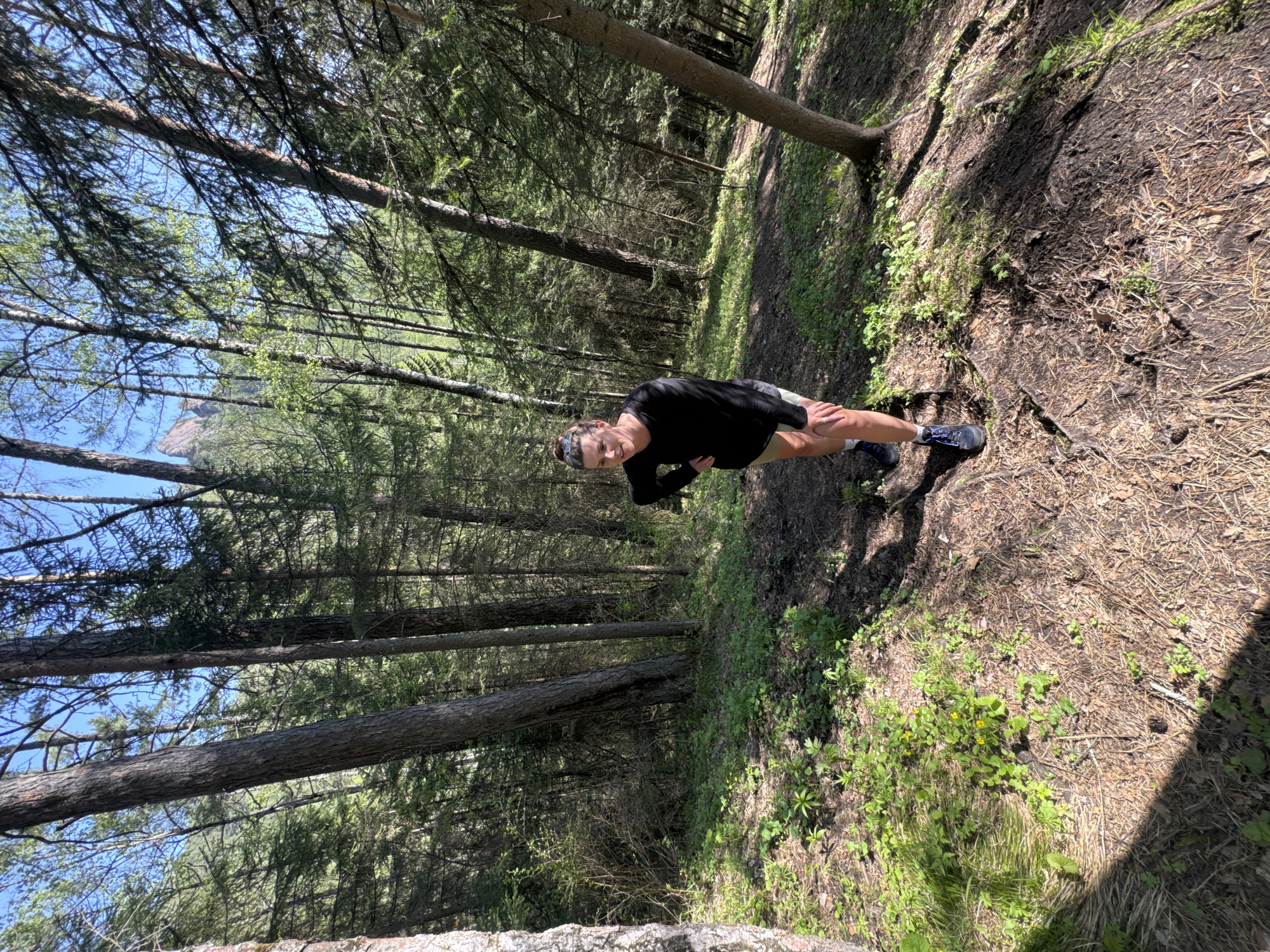 The image captures a woman in the Stolby Nature Reserve, located in Krasnoyarsk, Russia. She is positioned on a dirt and pine needle covered path within a dense forest of coniferous trees. The woman wears a black long-sleeved shirt, light-colored shorts, and athletic shoes. Her right hand is on her knee, and her left arm is bent with her hand near her side. The background features tall trees reaching up towards a partly visible blue sky. The photograph is taken at an angle. The path is covered in fallen pine needles and some sparse green vegetation. The lighting is a mix of sunlight and shadows, creating a dappled effect on the ground and trees.