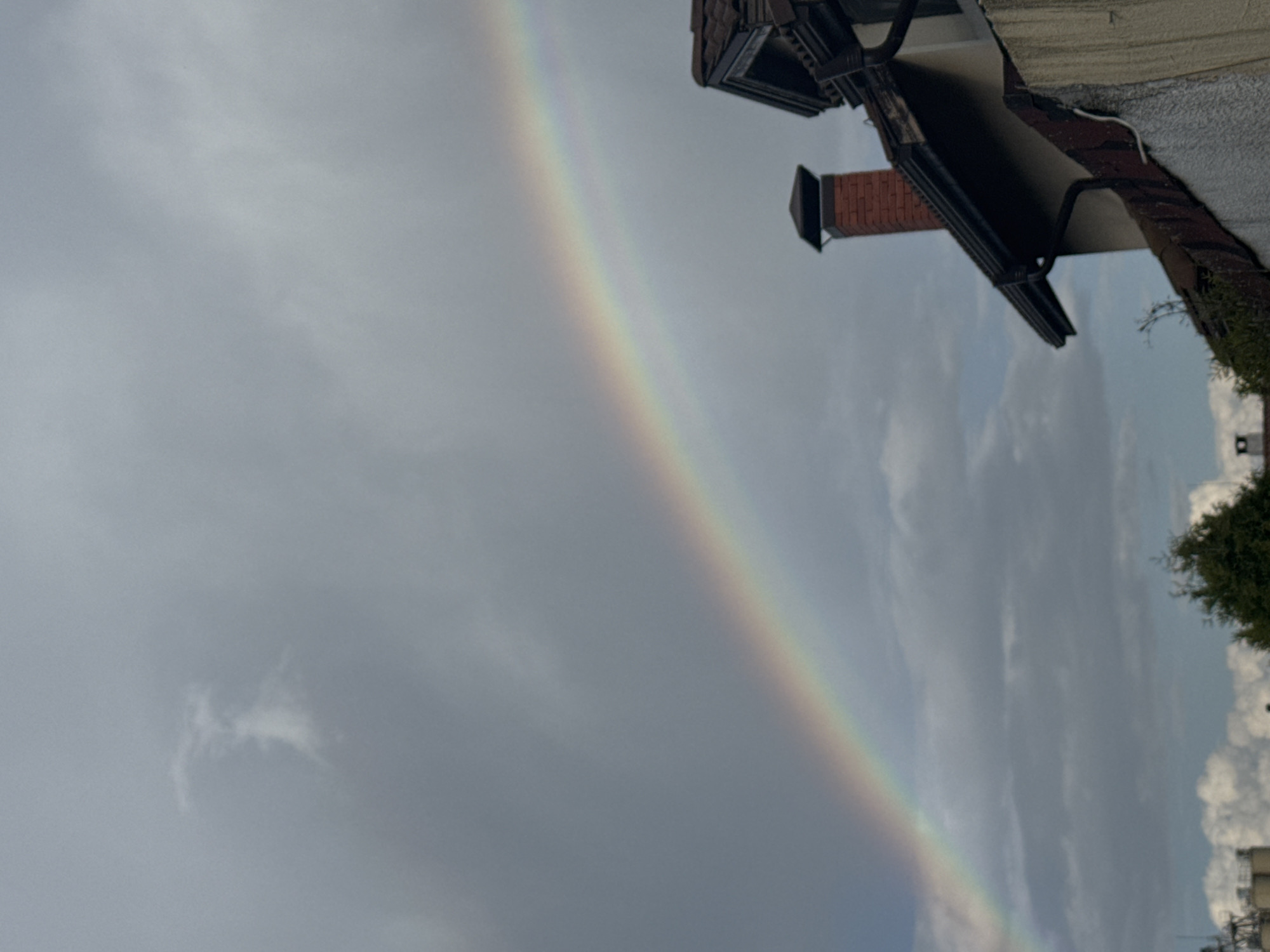 The image shows a rainbow in the sky above Bozhurishte, Bulgaria. A portion of a building, including a chimney and gutter, is visible in the upper right corner. Clouds fill the sky. Trees and other buildings are seen in the lower right portion of the image.