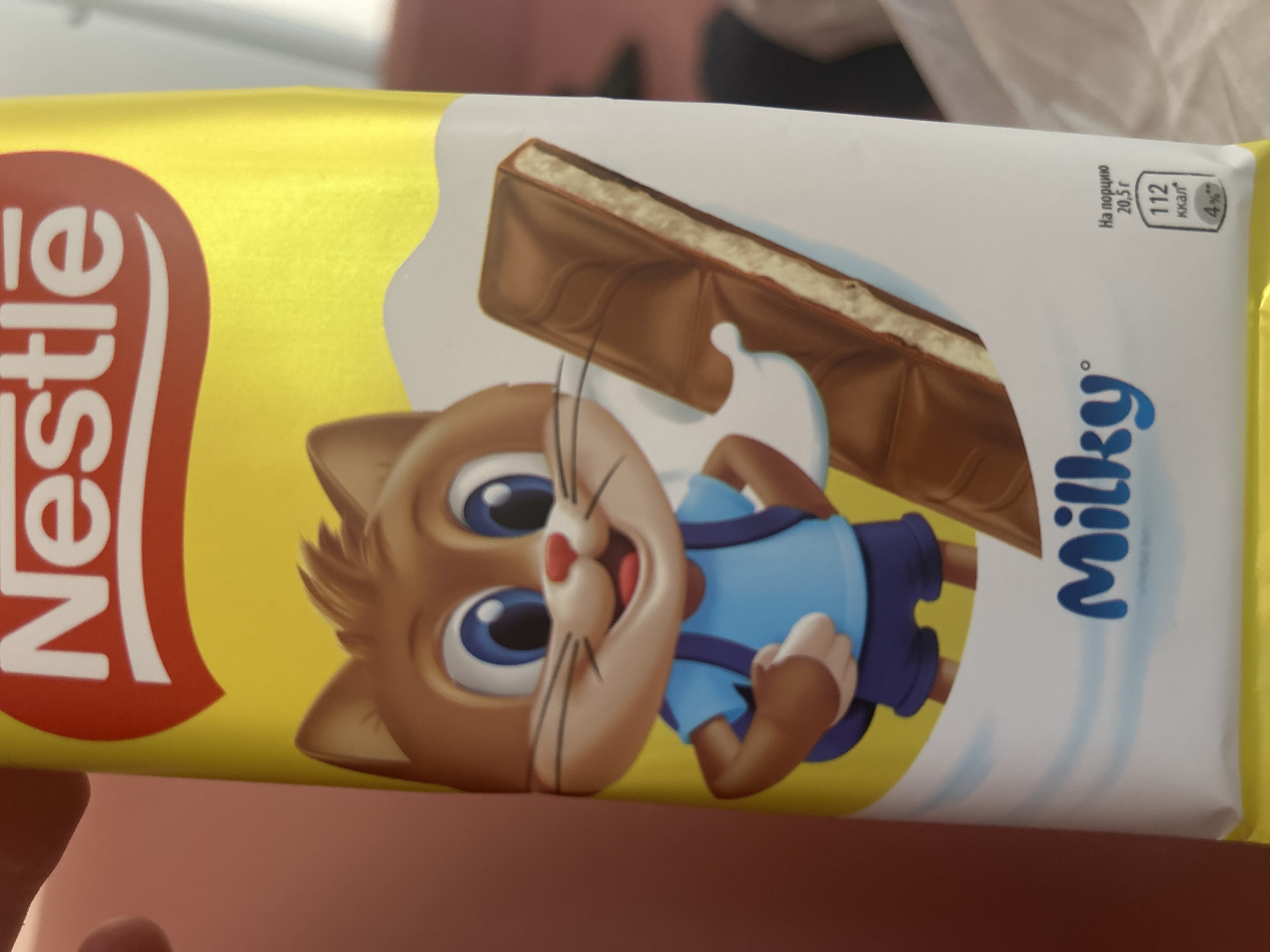 The image shows a close-up of a Nestle Milky chocolate bar packaging. The packaging is primarily yellow and features a cartoon brown cat wearing a blue t-shirt and purple shorts, carrying a blue backpack. A chocolate bar is depicted next to the cat, showing its layers. The text "Nestle" is visible at the top, and "Milky" is written in blue font at the bottom of the packaging. Nutritional information is also present, indicating "112 ккал" (112 kcal) per 20.5g serving, which is 4% of some unspecified daily value. The background is a blur of pink and white, suggesting an indoor setting, possibly a store or a home. There is no specific information in the image that allows for the determination of its location in Minsk, Belarus.