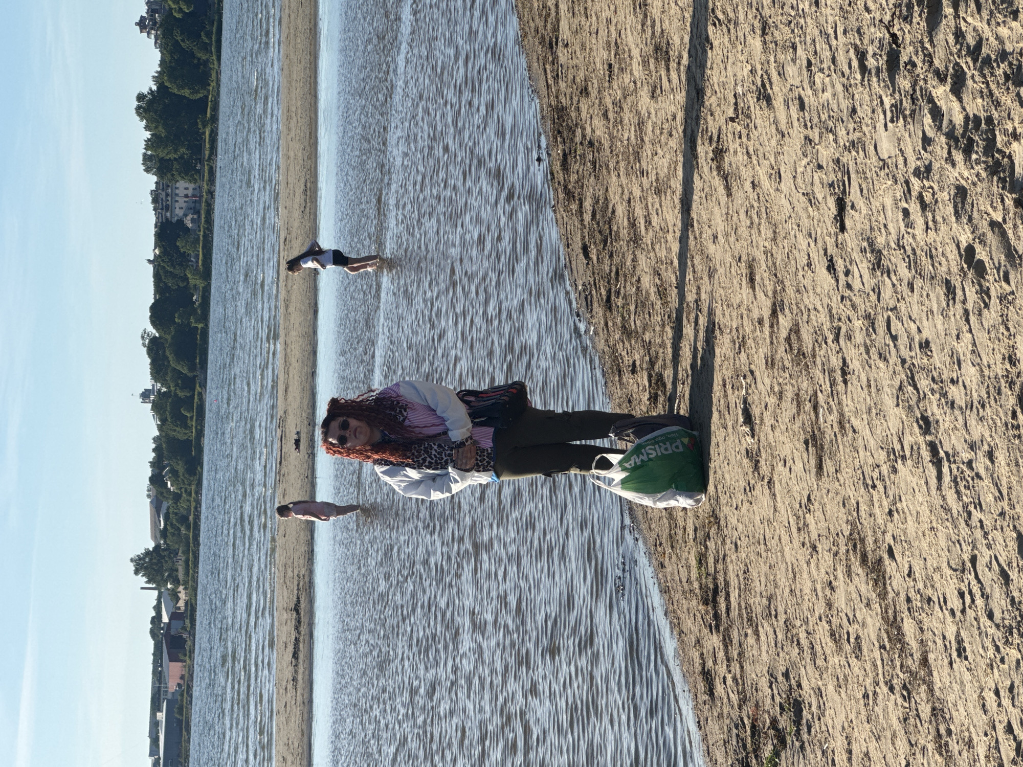 The image depicts a sunny day at a beach in Tallinn, Estonia. In the foreground, a person with bright red curly hair stands on a sandy shore, facing the camera. They are wearing sunglasses, a light-colored jacket with colorful stripes, a leopard-print scarf, and olive green pants. In front of them, there are two bags, one green with "PRISMA" written on it and another dark-colored one.
In the background, shallow water extends from the shore, with sandbars visible in the distance. Three other individuals are in the water: one person stands further back in the water, and two others, appearing to be children, are closer to the sandbar. Beyond the water, a cityscape with buildings and trees lines the horizon under a clear blue sky. The scene suggests a recreational setting, possibly a beach frequented by locals and visitors.