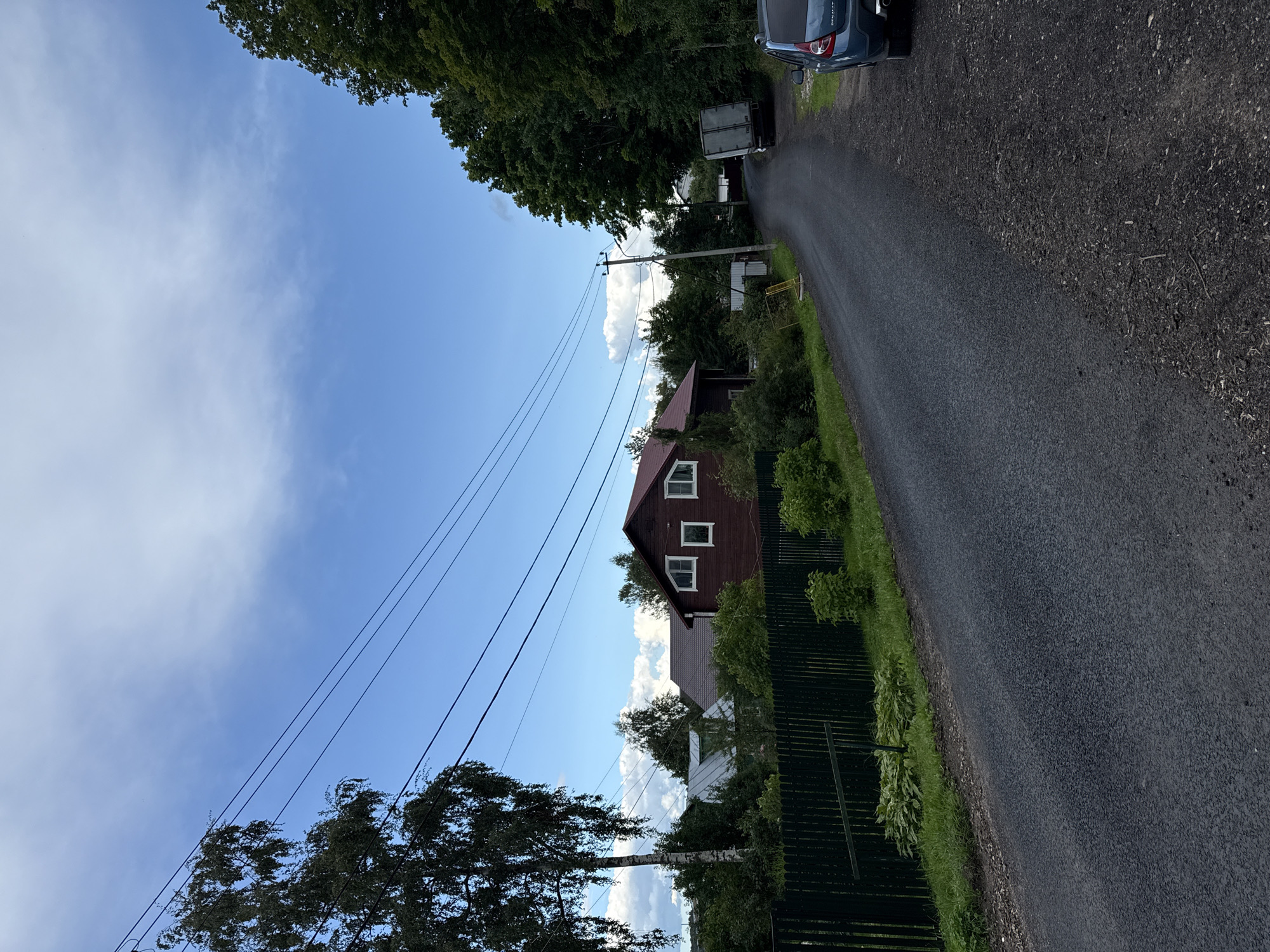 The image depicts a rural road in Krasnoye Selo, Russia, under a partly cloudy sky. On the left side of the road, a dark green wooden fence runs alongside a property with lush green vegetation and trees. A tall birch tree with dark bark and sparse leaves stands prominently on the left. Further back, a red wooden house with a white-trimmed roof and several white-framed windows is visible. Other houses and trees are scattered in the background. The road itself is paved and curves towards the right, leading into a dense forest on the right side of the image. A light-colored truck is parked on the right side of the road, and a dark blue car is parked further down the road. Electrical wires are strung across the sky, supported by utility poles. The overall scene suggests a quiet, residential area in a natural setting.