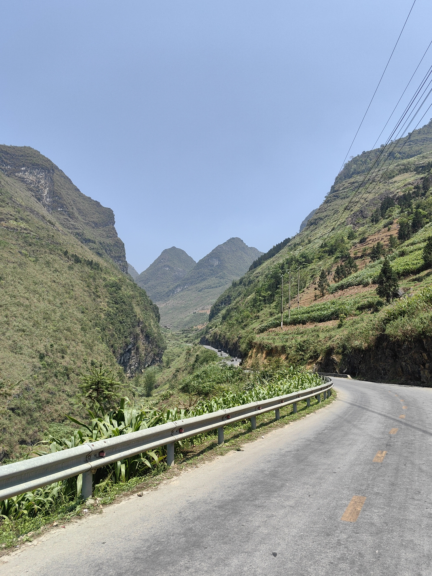 The image shows a road winding through a mountainous region in Phố Bảng Commune, Vietnam. The road is paved and has a metal guardrail along one side. Vegetation covers the slopes of the mountains. Power lines run overhead. The sky is clear and blue.