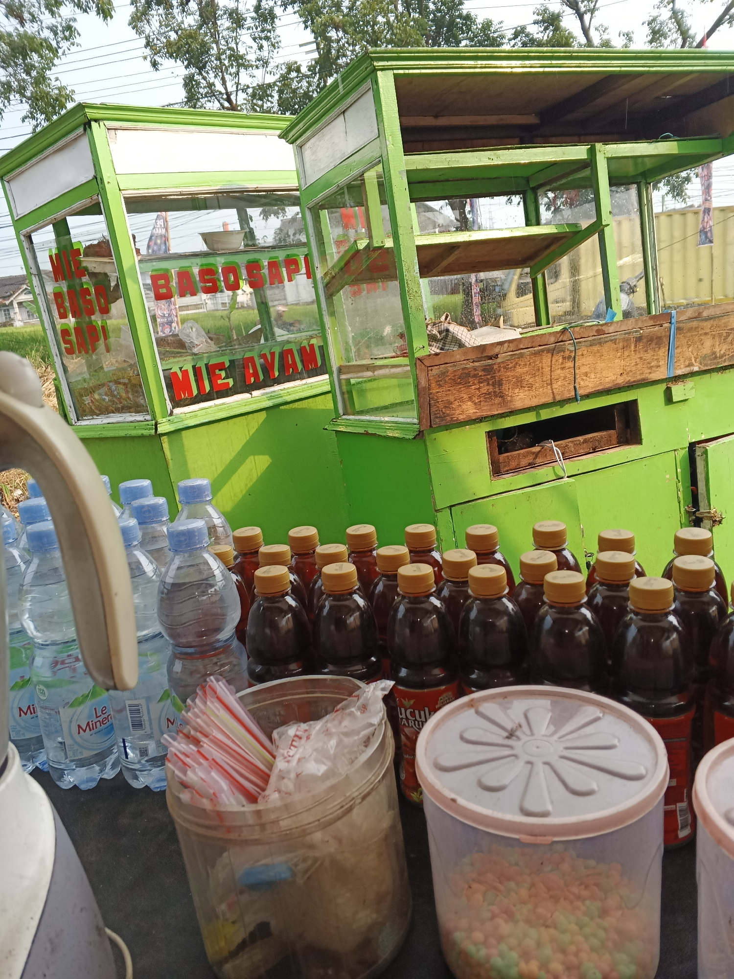 The image captures a street-side food stall in Sukabumi, Indonesia, likely specializing in "Mie Ayam" (chicken noodles) and "Baso Sapi" (beef meatballs), as indicated by the signage on the green carts. Two mobile food carts, painted bright green with white trim and glass display cases, are positioned side-by-side. The cart in the foreground displays "MIE BASO SAPI" and "MIE AYAM" in red lettering.
In front of the carts, a variety of items are laid out on a dark surface. These include several plastic bottles of "Le Minerale" water, a container filled with red and white striped straws, and a larger clear plastic container holding colorful, small, bead-like food items, possibly candies or snacks. Behind these items, rows of dark-colored bottled drinks with yellow caps are visible.
The environment suggests an outdoor setting with trees and foliage in the background, bathed in natural sunlight, indicating daytime. The presence of buildings in the distance suggests an urban or semi-urban location within Sukabumi. No people are clearly visible or actively engaged in transactions in the immediate foreground, though the setup implies a food vending operation. The overall scene is one of a typical Indonesian street food stall ready for customers.