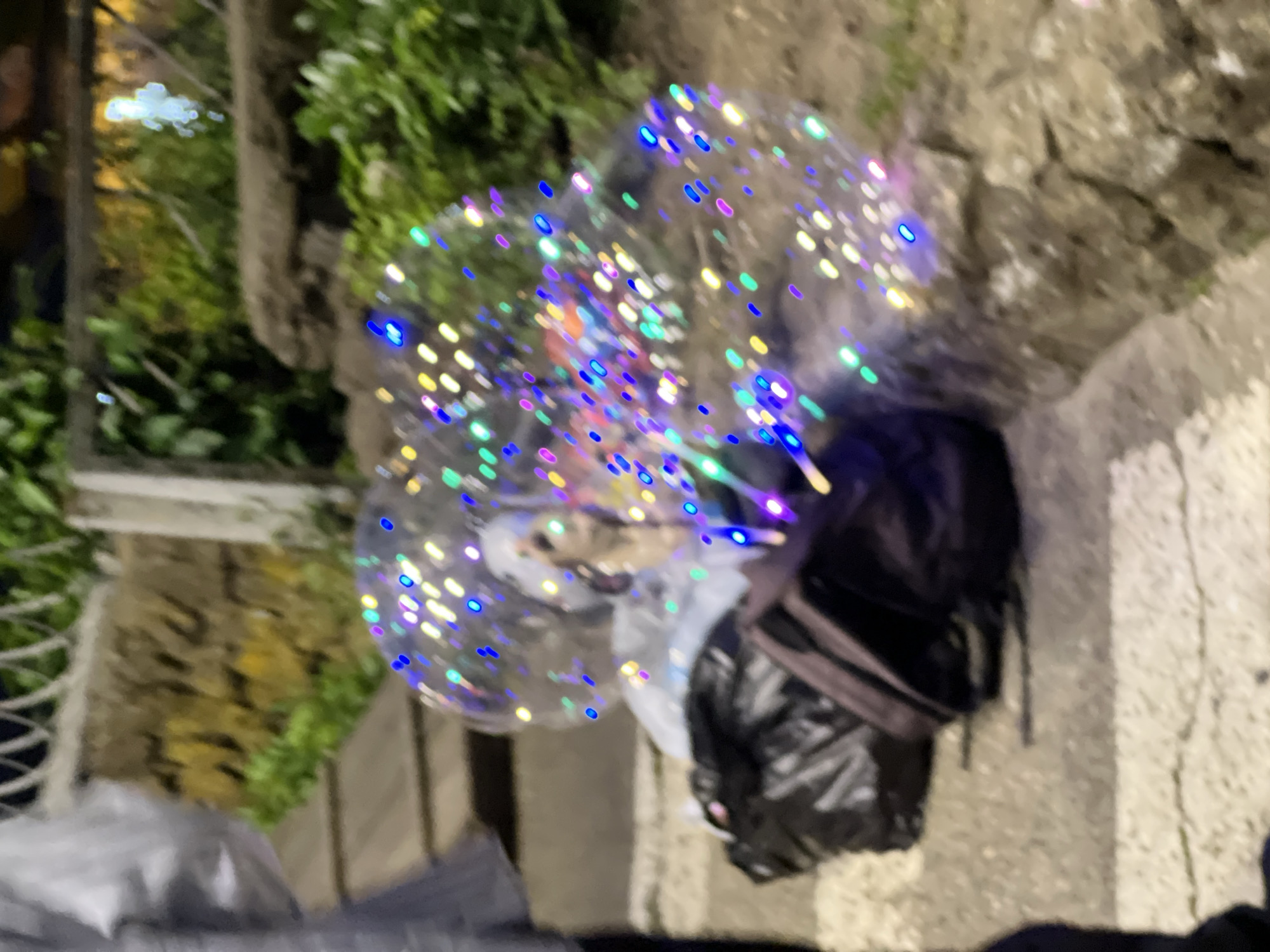 The image shows a person, partially visible on the left, wearing a light-colored shirt. In the center of the image, a person is seated, covered by a clear balloon filled with colorful LED lights. They are surrounded by several bags, including a dark backpack and a black trash bag, on a paved surface that appears to be a sidewalk or walkway. The background consists of a low stone wall, greenery, and what looks like a fence. The scene is dimly lit, suggesting it's evening or nighttime, and the LED lights in the balloon are the primary light source, casting a festive glow. There is no specific information in the image to determine the location in Krasnodar, Russia.