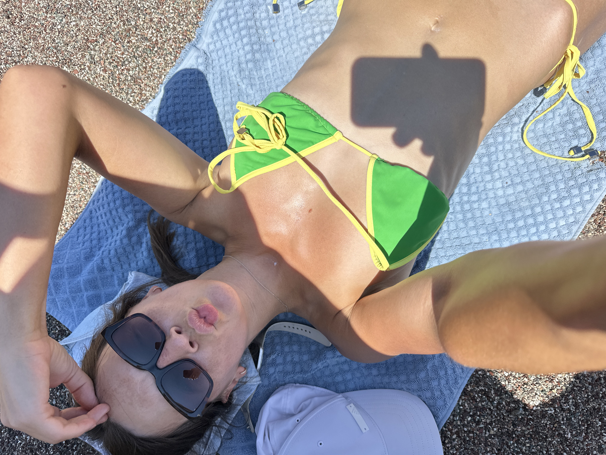 A young woman is lying on a blue towel on a beach in Boreti, Montenegro. She is wearing a green and yellow bikini and sunglasses. Her right hand is resting on her head, and her left arm is extended towards the camera. The beach is covered in small pebbles. A white baseball cap and a watch are lying on the towel next to her. The sun is shining brightly, casting shadows on her body and the towel. She is making a kissing face at the camera.