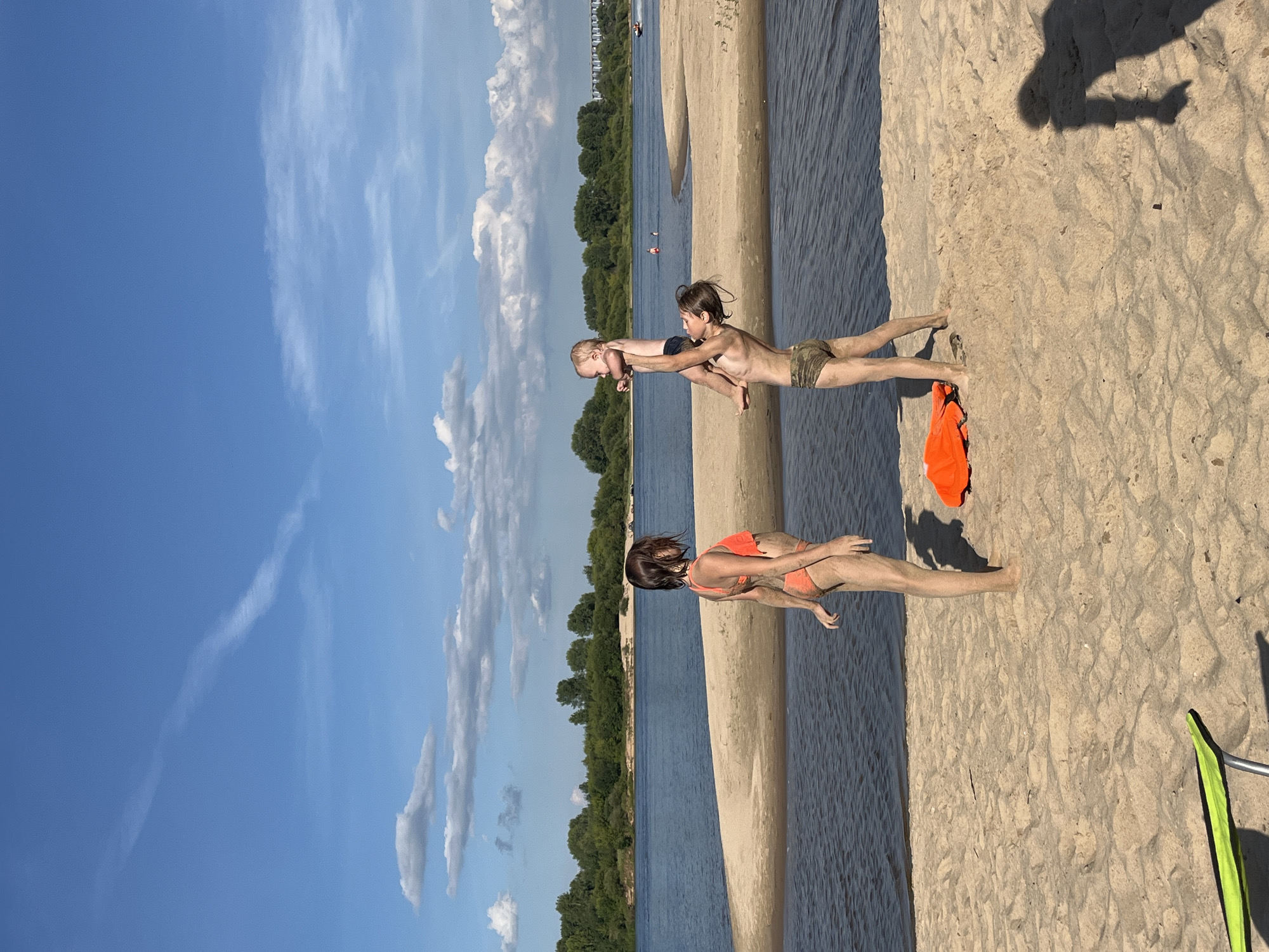 The image depicts a bright, sunny day on a sandy beach with a body of water in the background, likely the Oka River in Nizhny Novgorod, Russia. The sky is a vibrant blue, dotted with scattered white clouds, suggesting fair weather. The scene is oriented vertically, with the sky occupying the left half of the frame and the beach and water on the right.
In the foreground, a woman in an orange bikini is sitting on the sand. Further back, a boy in camouflage shorts is holding a baby, lifting it in the air. The boy appears to be playing with the baby, who is looking towards the boy. The overall mood seems to be one of playful family interaction on a warm summer day.
Notable details include the wet sand near the water's edge, indicating that the tide might have recently receded or that the water level is high. An orange life jacket is lying on the sand near the woman, and a green object, possibly a part of beach equipment, is visible in the lower right corner. In the distance, a treeline borders the far bank of the river, with some buildings visible along the horizon. The sunlight creates distinct shadows on the sand, suggesting it is likely midday or early afternoon. No visible text is present in the image.